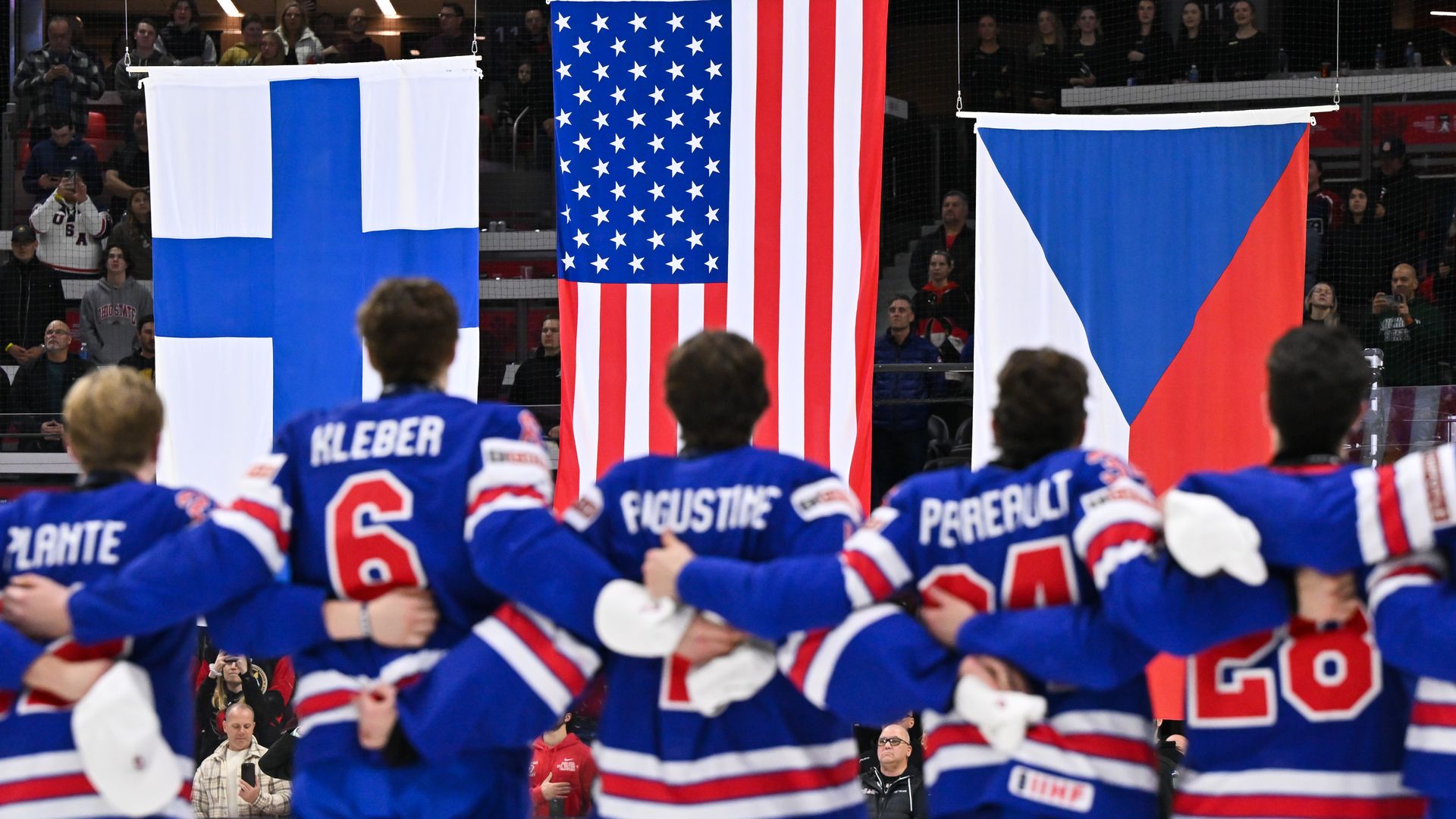 Hockey players in blue jerseys stand arm-in-arm facing flags of Finland, USA, and Czech Republic during a ceremony in an arena with spectators in the background.