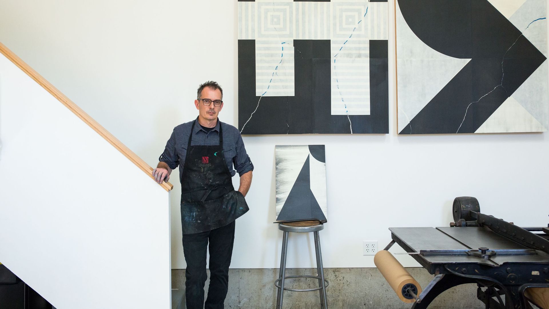 A man in a black apron standing in front of two paintings and a letter press