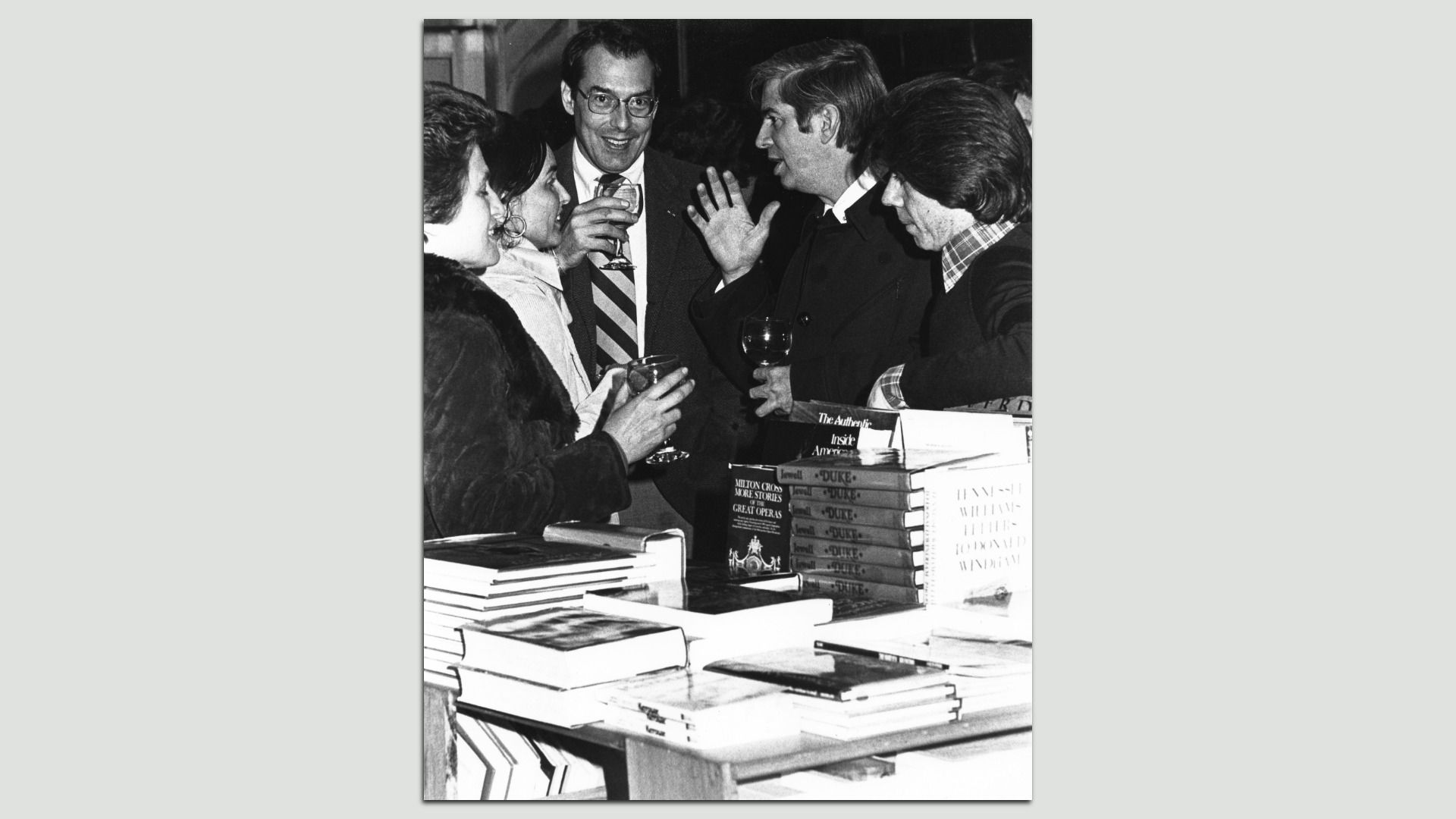 Black-and-white photo of five people at a book event, center man with glasses and a striped tie holding a drink, others converse with wine glasses; stacks of books on a table.