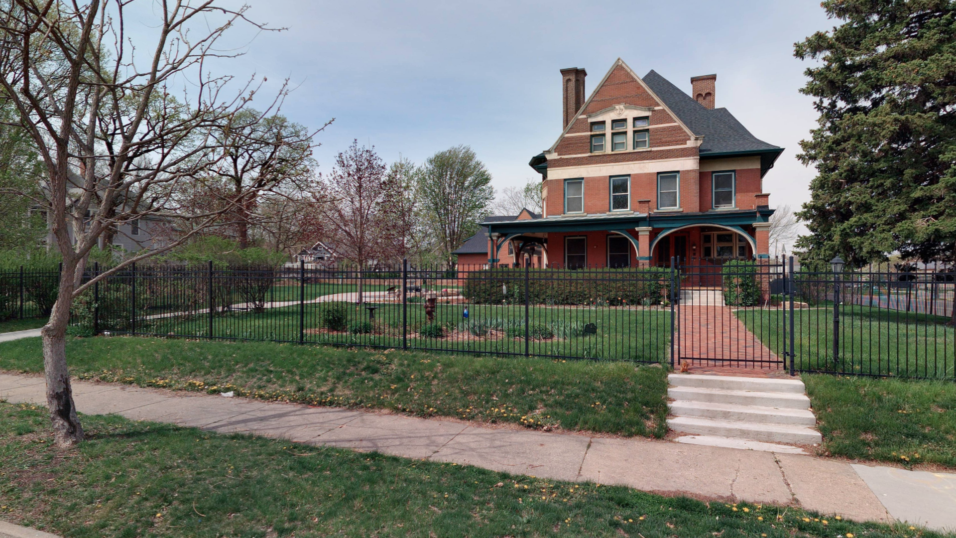 A large red brick Victorian house with a peaked roof, multiple chimneys, and a blue-green wraparound porch, enclosed by a black metal fence; a brick path leads to front steps across a green lawn.