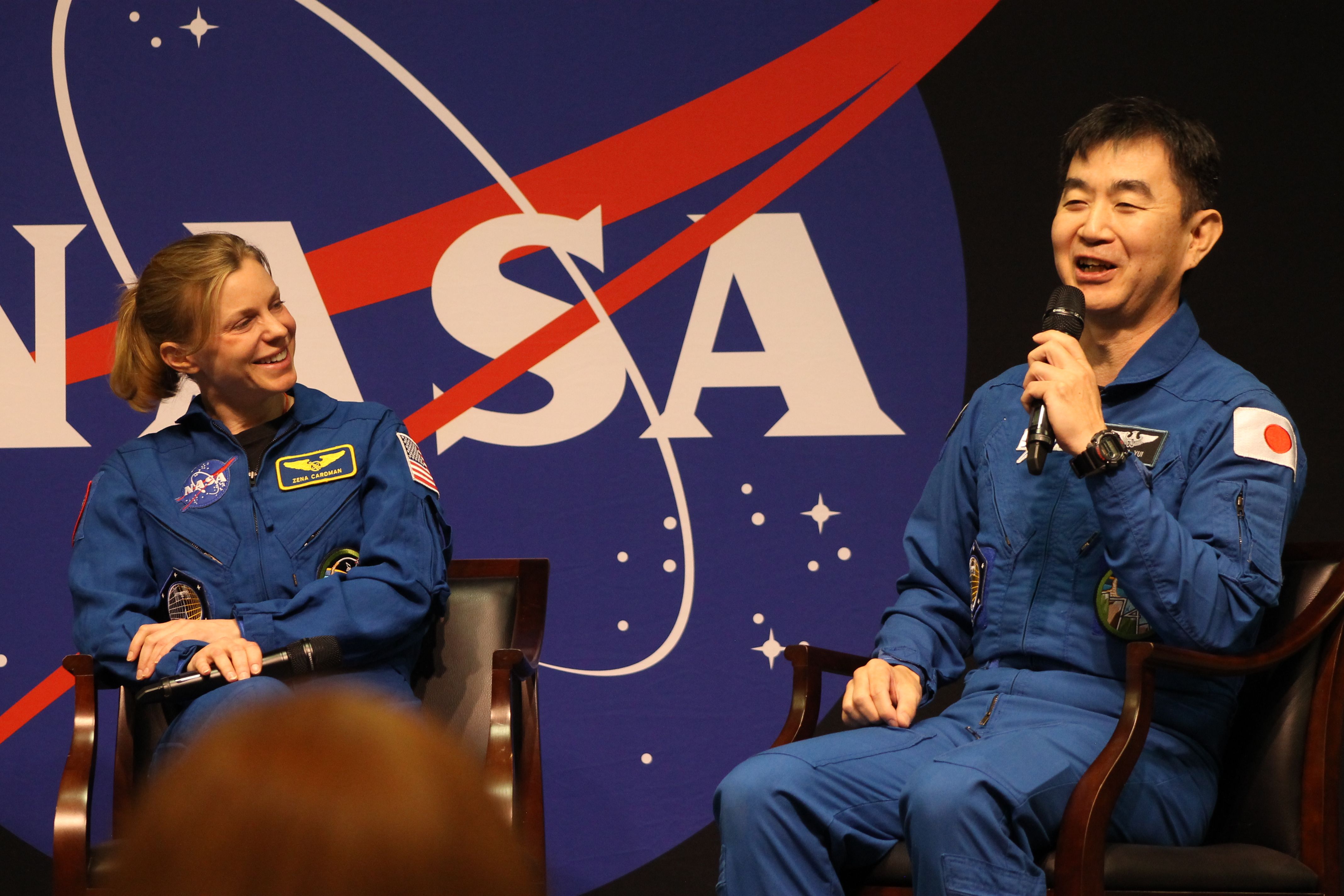 Two astronauts in blue NASA jumpsuits sit on stage in front of a large NASA backdrop; the woman on the left smiles with a mic, while the man on the right speaks into a handheld microphone.