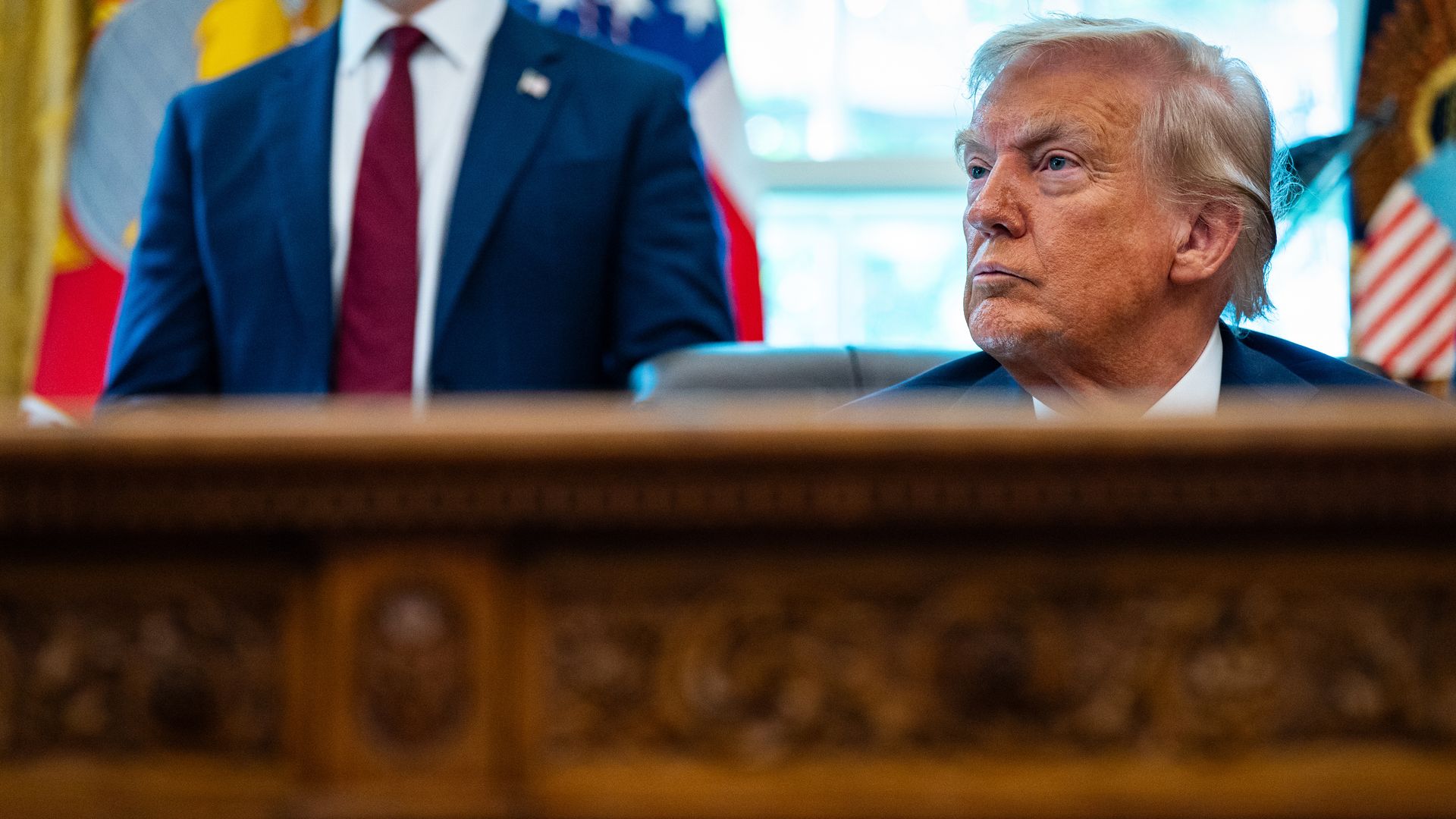 Former President Donald Trump sitting at an ornate wooden desk, looking right, with a person standing behind him wearing a blue suit and red tie, and flags blurred in the background.