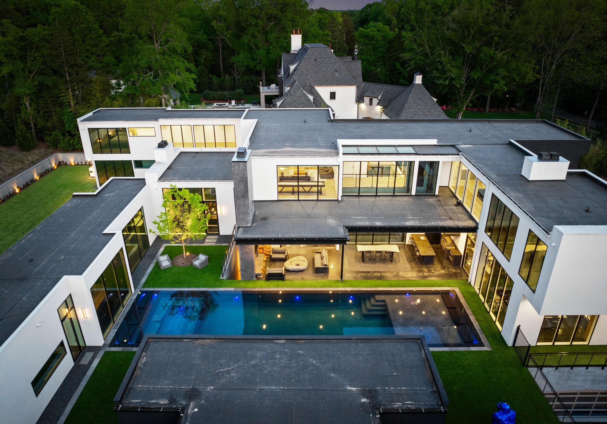 Aerial view of a modern white two-story house with flat roofs, large windows, outdoor seating, green lawn, and a rectangular blue-lit swimming pool at twilight.