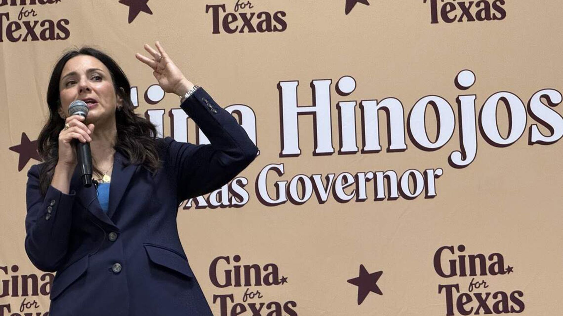 Woman speaks into a microphone on a beige backdrop with "Gina for Texas" branding and stars; wearing a navy blazer and blue pants, gesturing with her left hand.