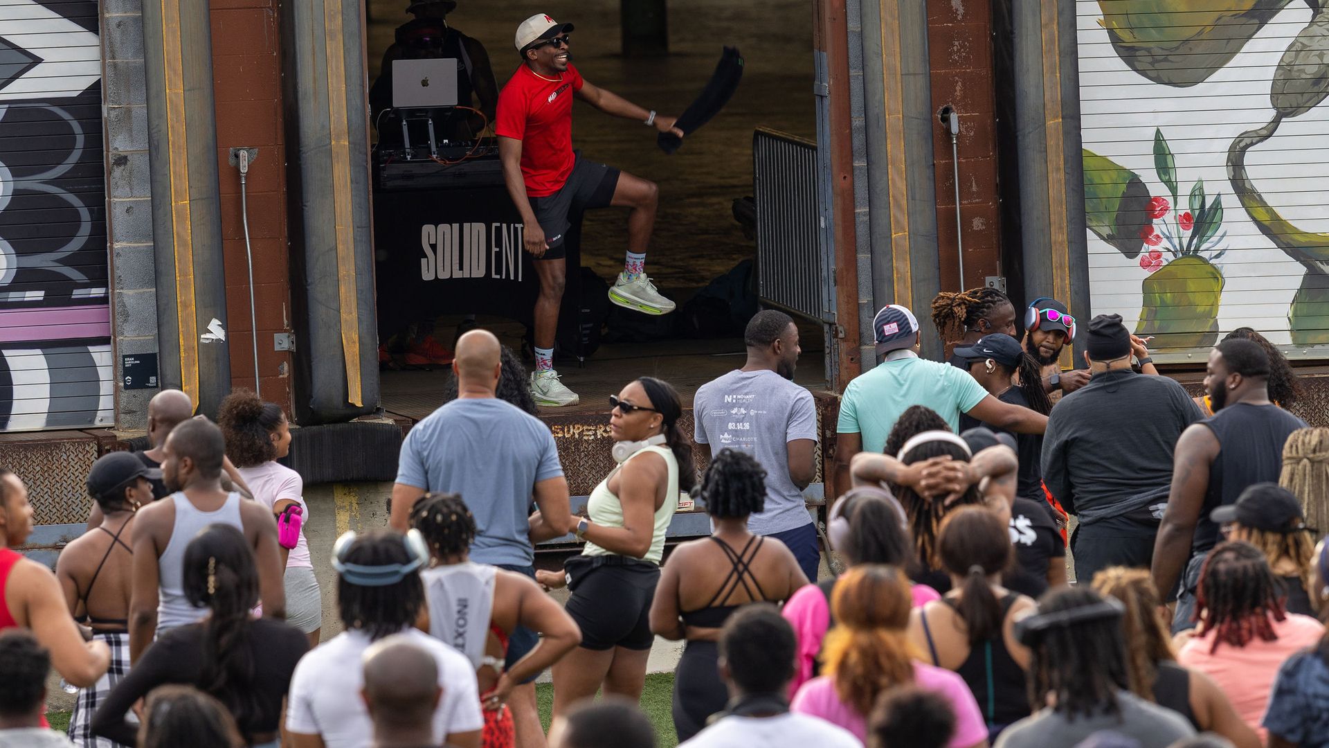 Crowded outdoor event outside a rusted brick warehouse. A man in a red shirt and white cap steps onto a platform near an open doorway with a DJ at a laptop; string lights hang overhead and a mural is on the right wall.