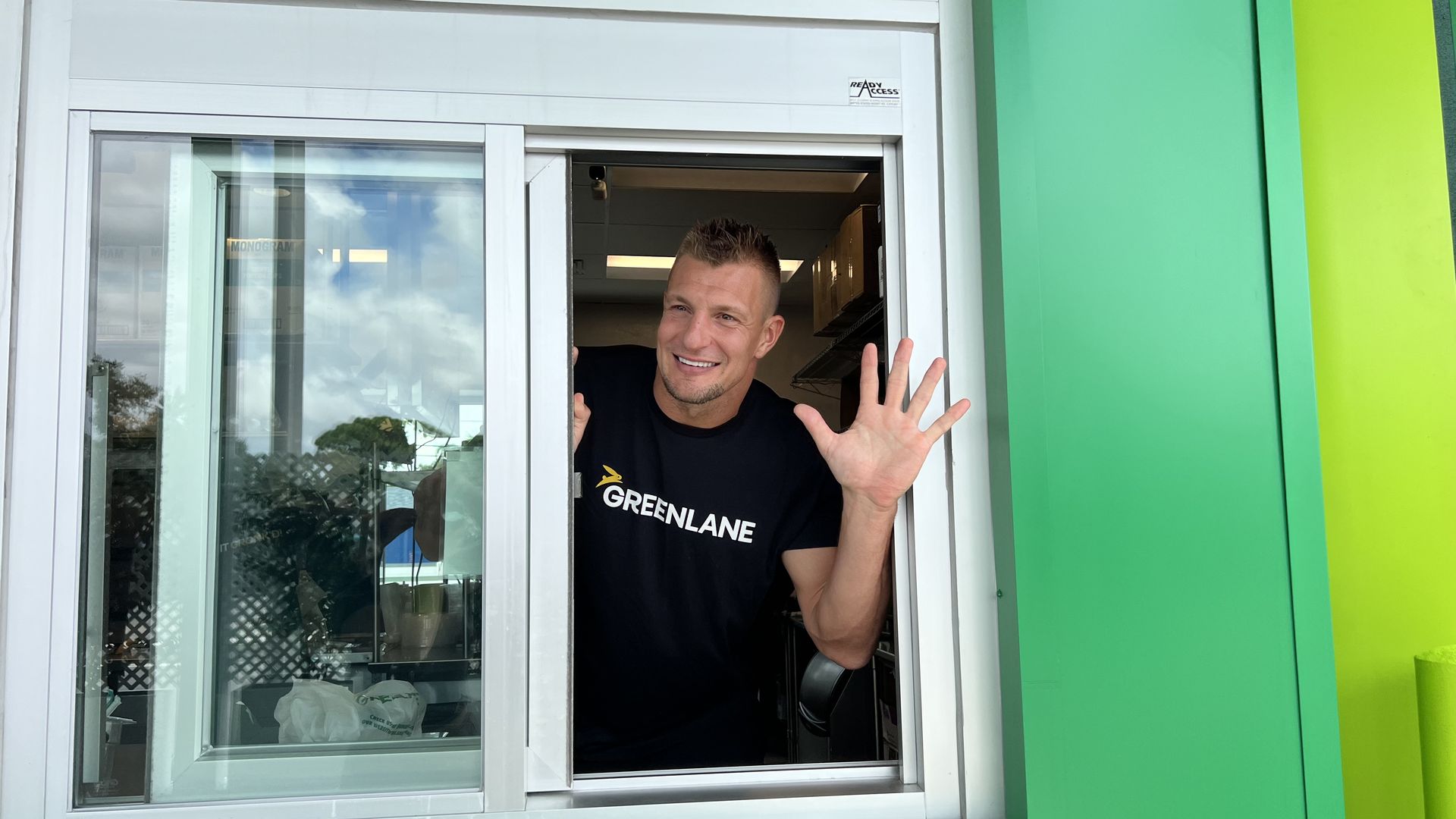 A man in a black T-shirt that says "GREENLANE" in white letters waving from a drive-thru window