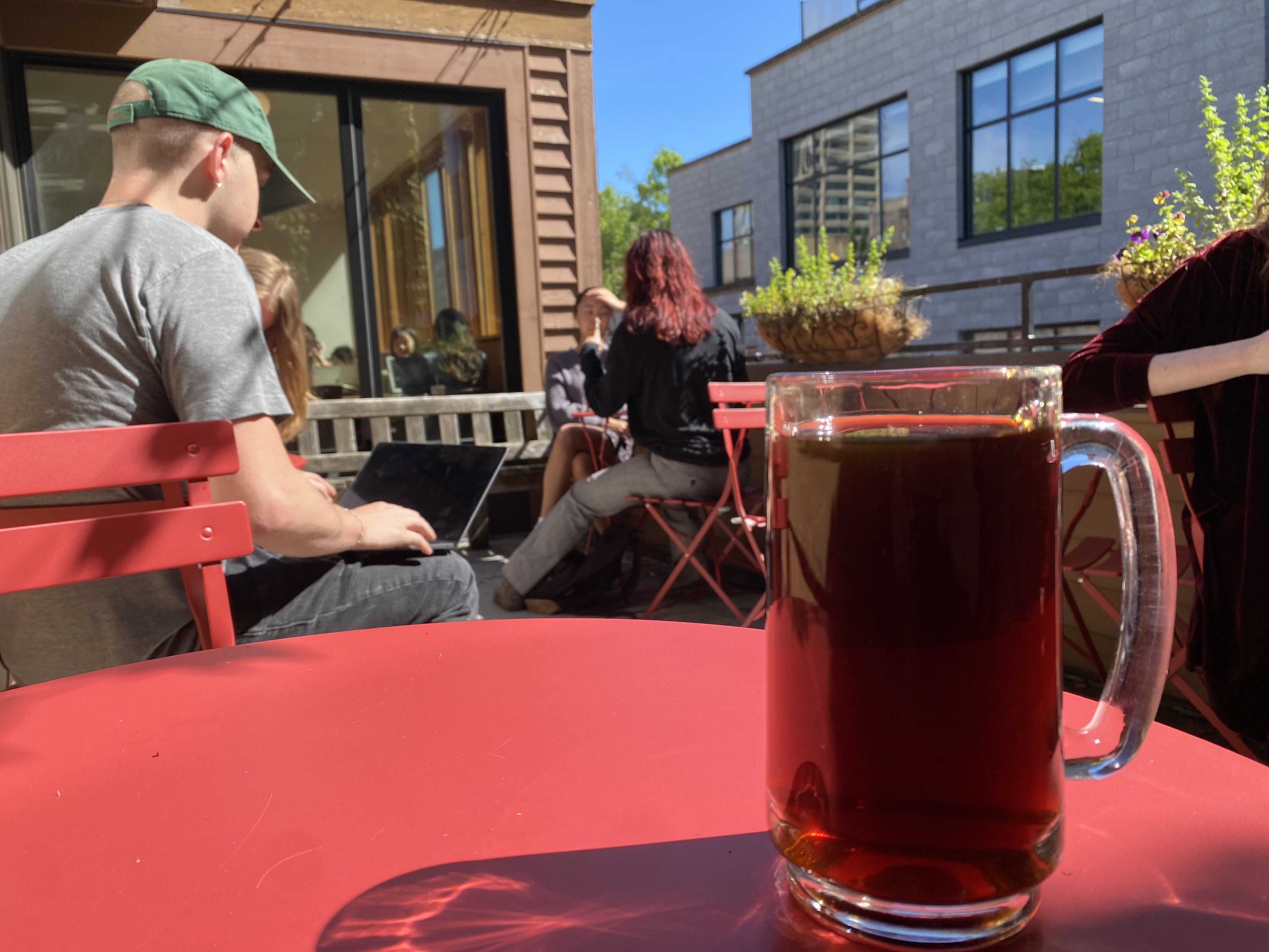 A glass mug full of black coffee sitting on an outdoor red table, with people at tables in the background and some plants on the edge of a wall.