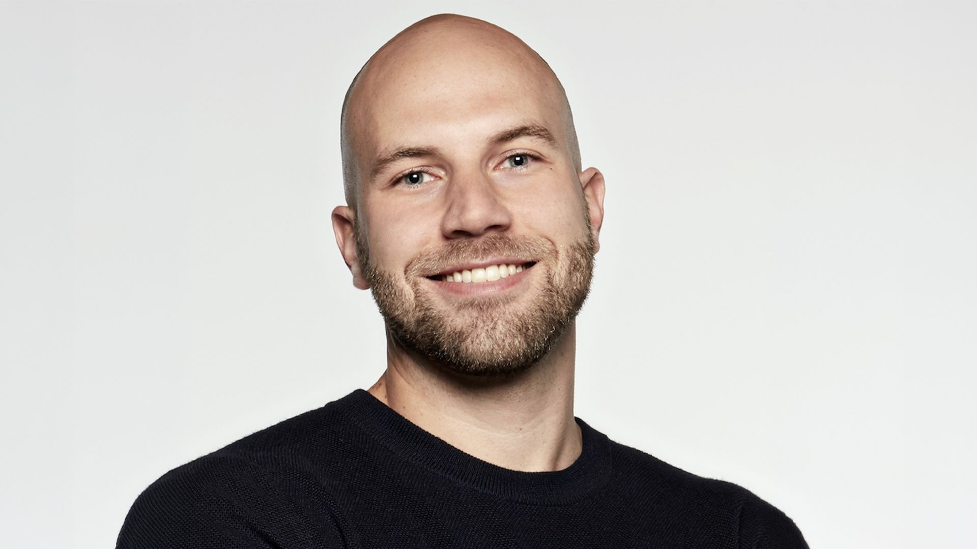 Smiling bald man with a short beard, wearing a dark navy sweater, posed against a pale gray background.