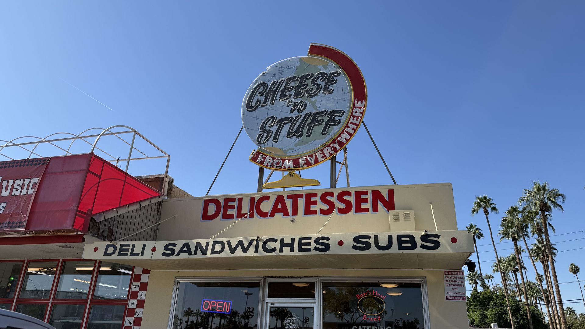 Exterior of a delicatessen with a large round sign saying "Cheese 'n Stuff From Everywhere". Below, a banner reads "Deli Sandwiches Subs". A neon "open" sign is illuminated in the window. 