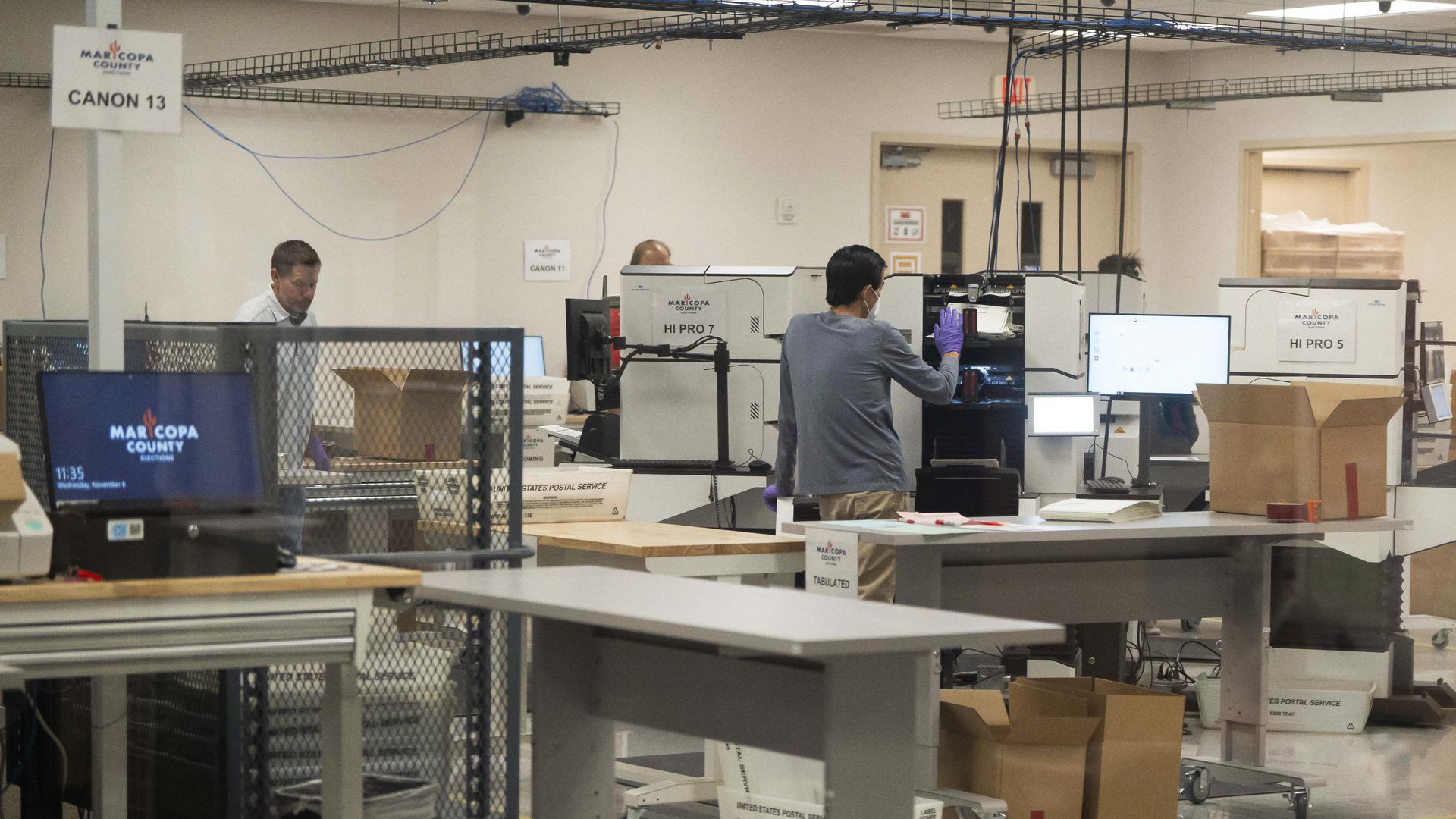 Inside a Maricopa County election processing facility, workers wearing masks and gloves operate ballot tabulation machines labeled HI PRO 5 and HI PRO 7 amid tables, boxes, and computers.