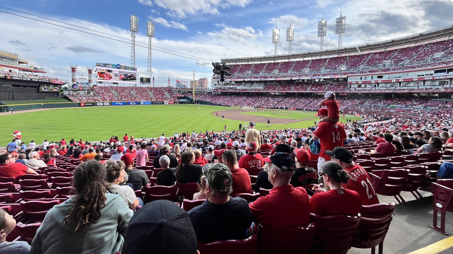Wide view of a baseball stadium filled with red-clad fans, bright green field, large floodlight towers, blue sky with scattered clouds, scoreboard and seating on left and right.
