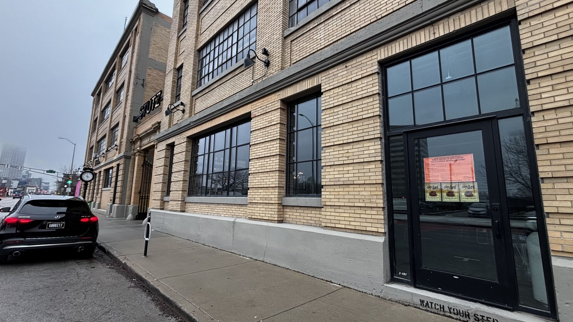 Exterior of a beige brick building with large black-framed windows and a black door displaying orange and yellow notices, a parked black car on a gray sidewalk under overcast sky.