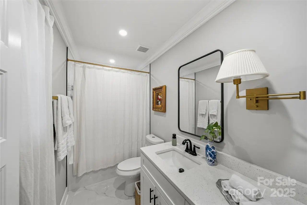 Modern bathroom with white walls, shower curtain, black-framed mirror, white vanity, gold wall lamp, towels, and blue-and-white vase with green plant.
