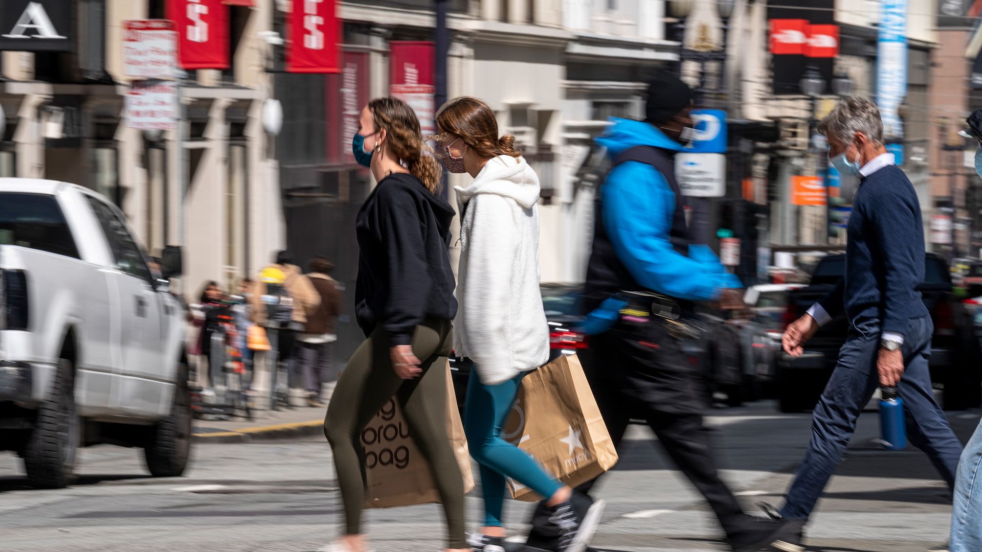 Shoppers wearing protective masks carry bags cross a street