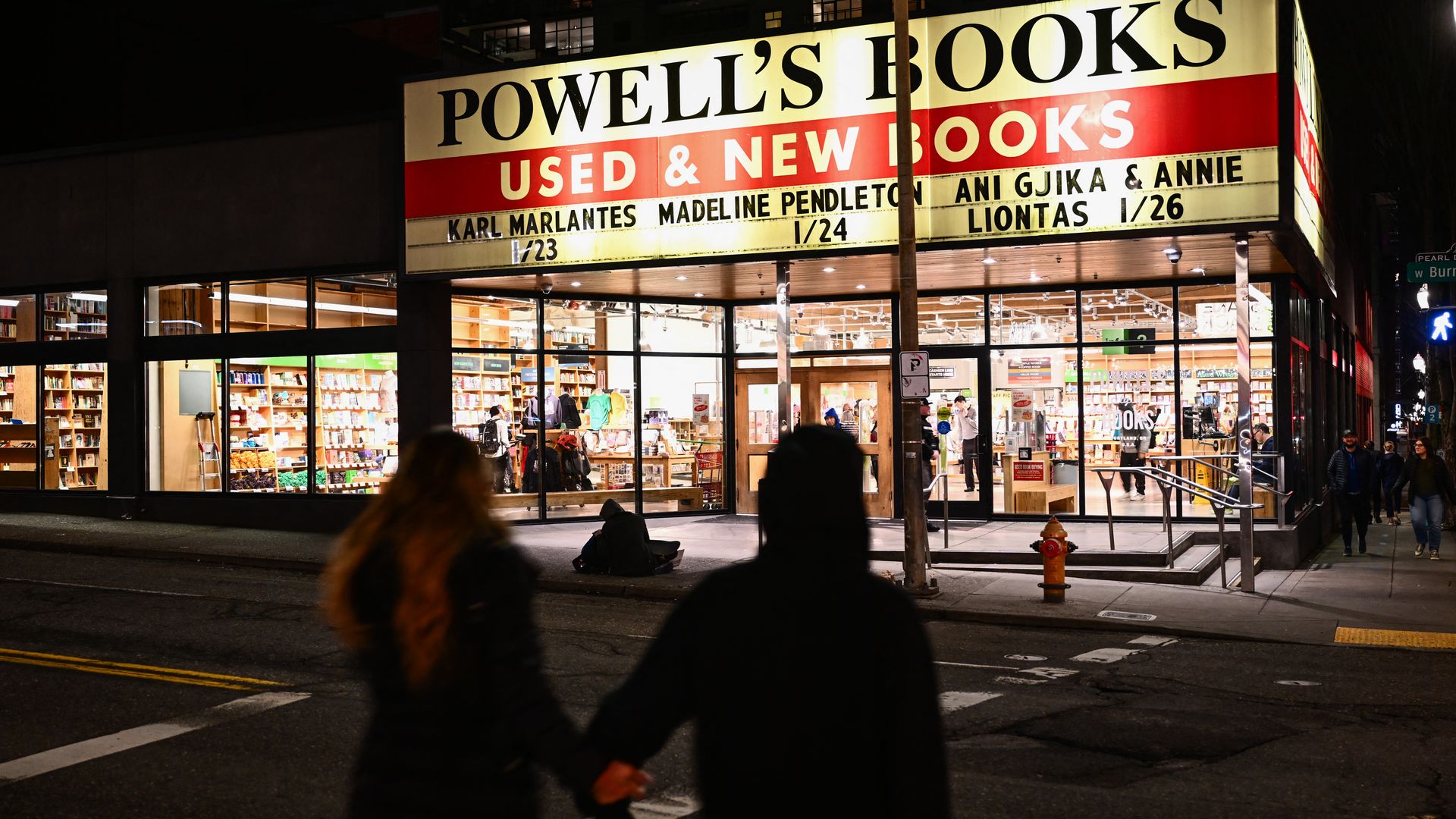 Two people hold hands and walk toward a building that has a sign that reads "Powell's Books"