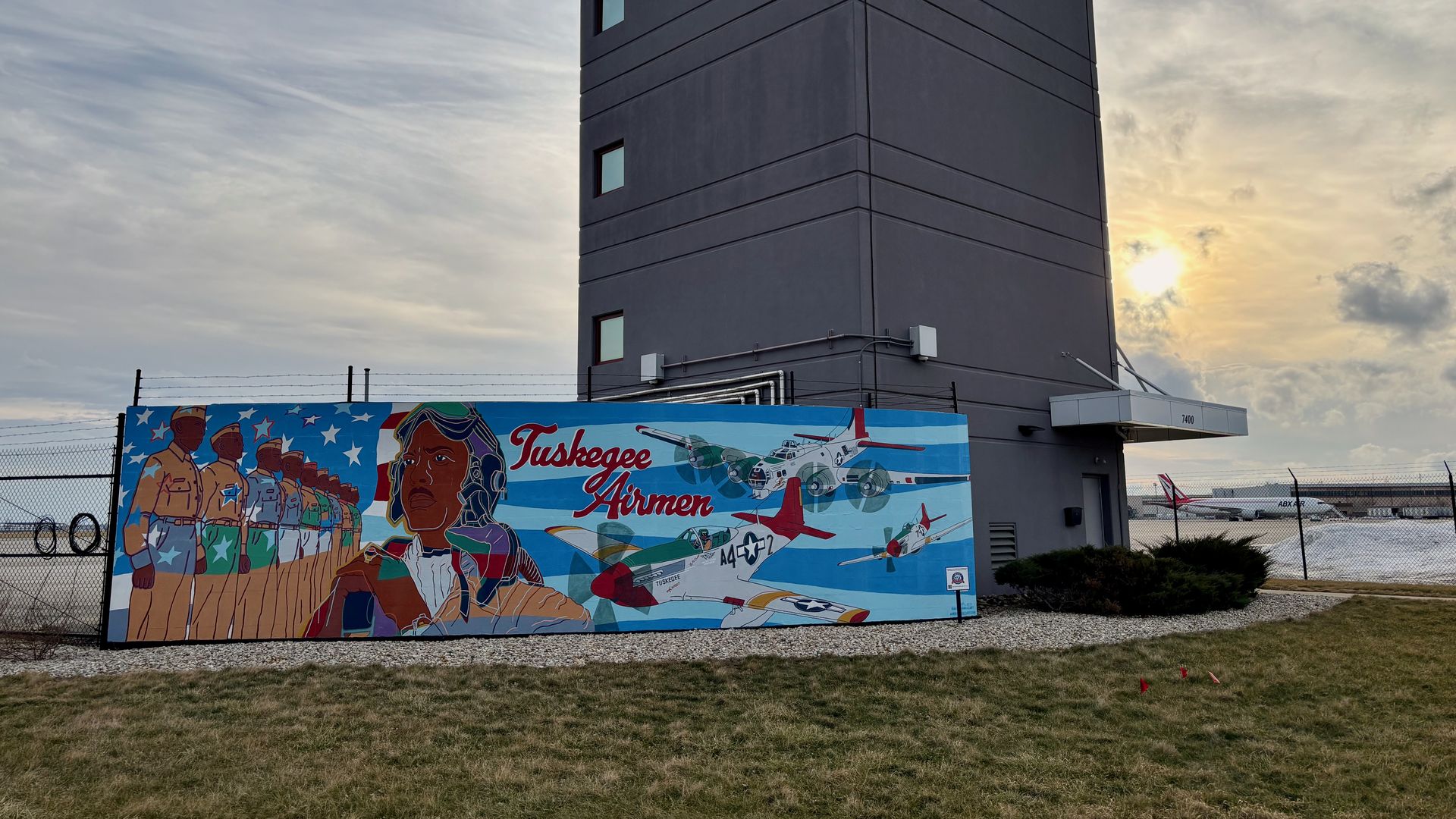 Colorful mural near gray tower shows Tuskegee Airmen in brown uniforms, vintage planes with red and white accents, and text "Tuskegee Airmen" on blue sky background at airport in sunset light.