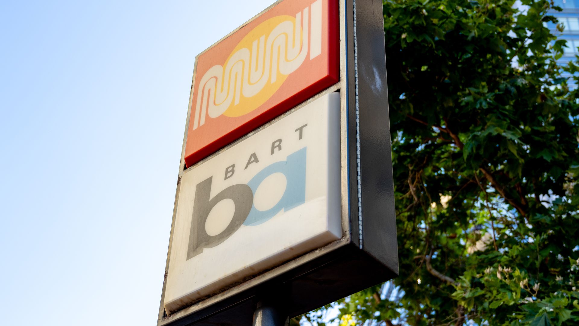 Two stacked transit signs against a blue sky and green tree background. The top sign is orange with a yellow circle and white stylized MUNI text. Below, a white BART logo with black and blue letters.
