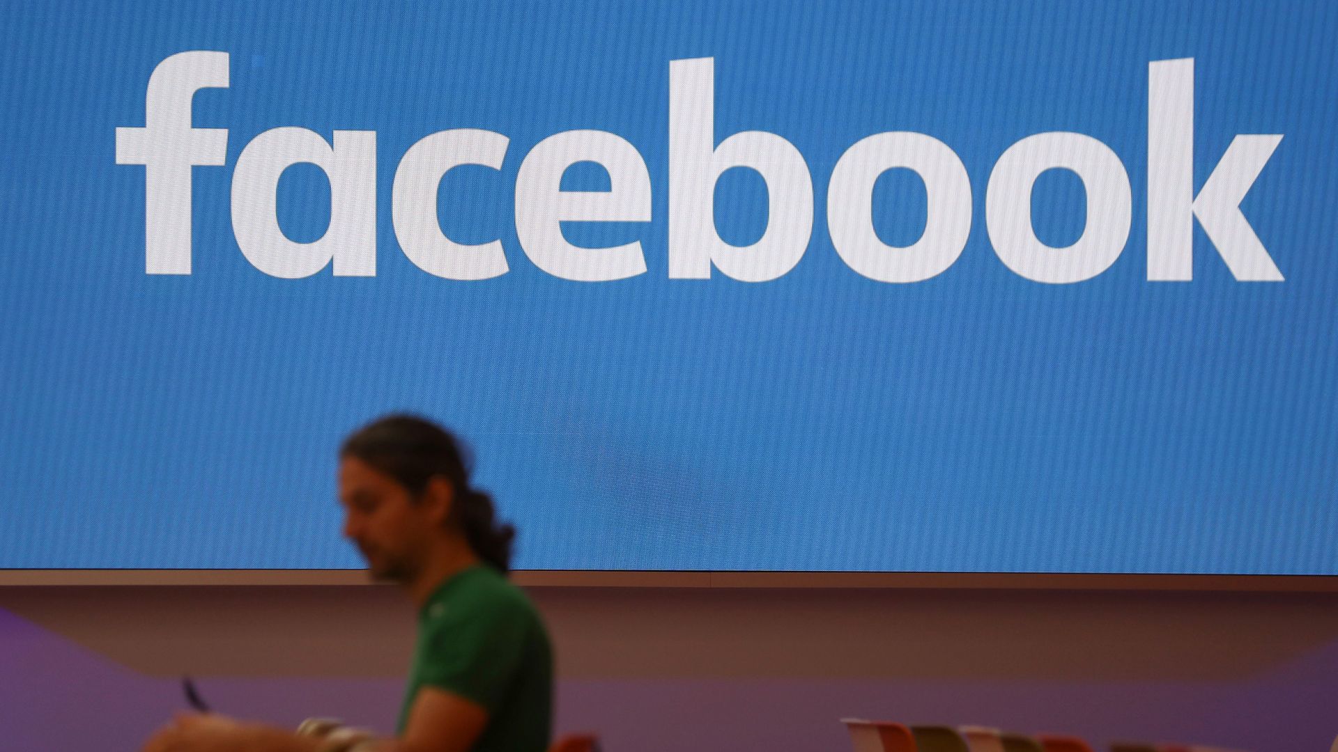 A man wearing a green t-shirt sits in front of a Facebook sign