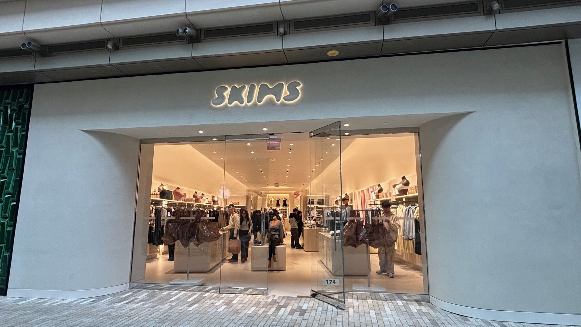 Front of a SKIMS store with glass doors, curved white facade, illuminated SKIMS sign, shoppers inside browsing clothing on racks under warm showroom lighting.