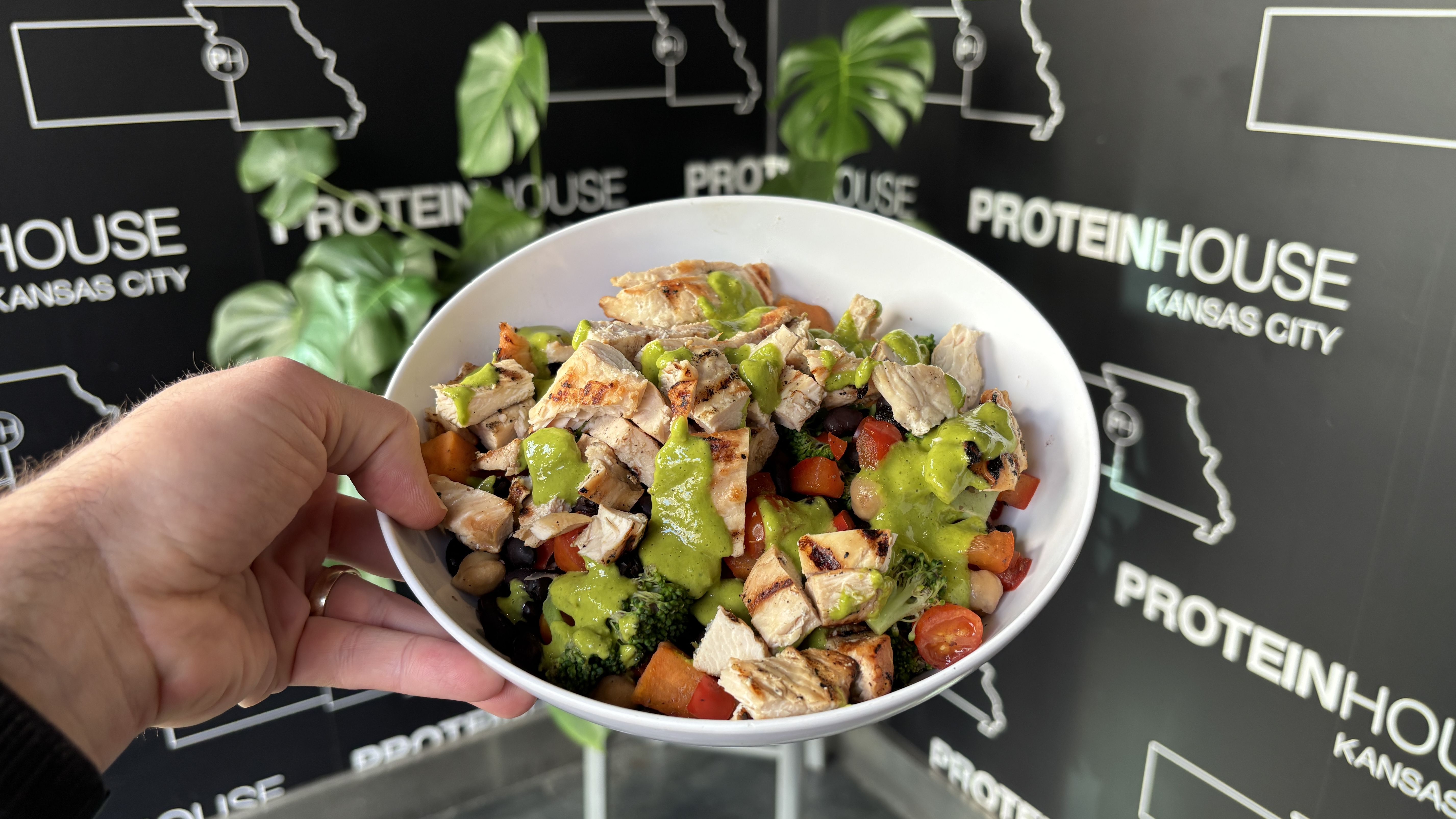 Hand holding white bowl with grilled chicken, broccoli, black beans, chickpeas, tomatoes, and green sauce, in front of a black Protein House Kansas City backdrop with green leaves.
