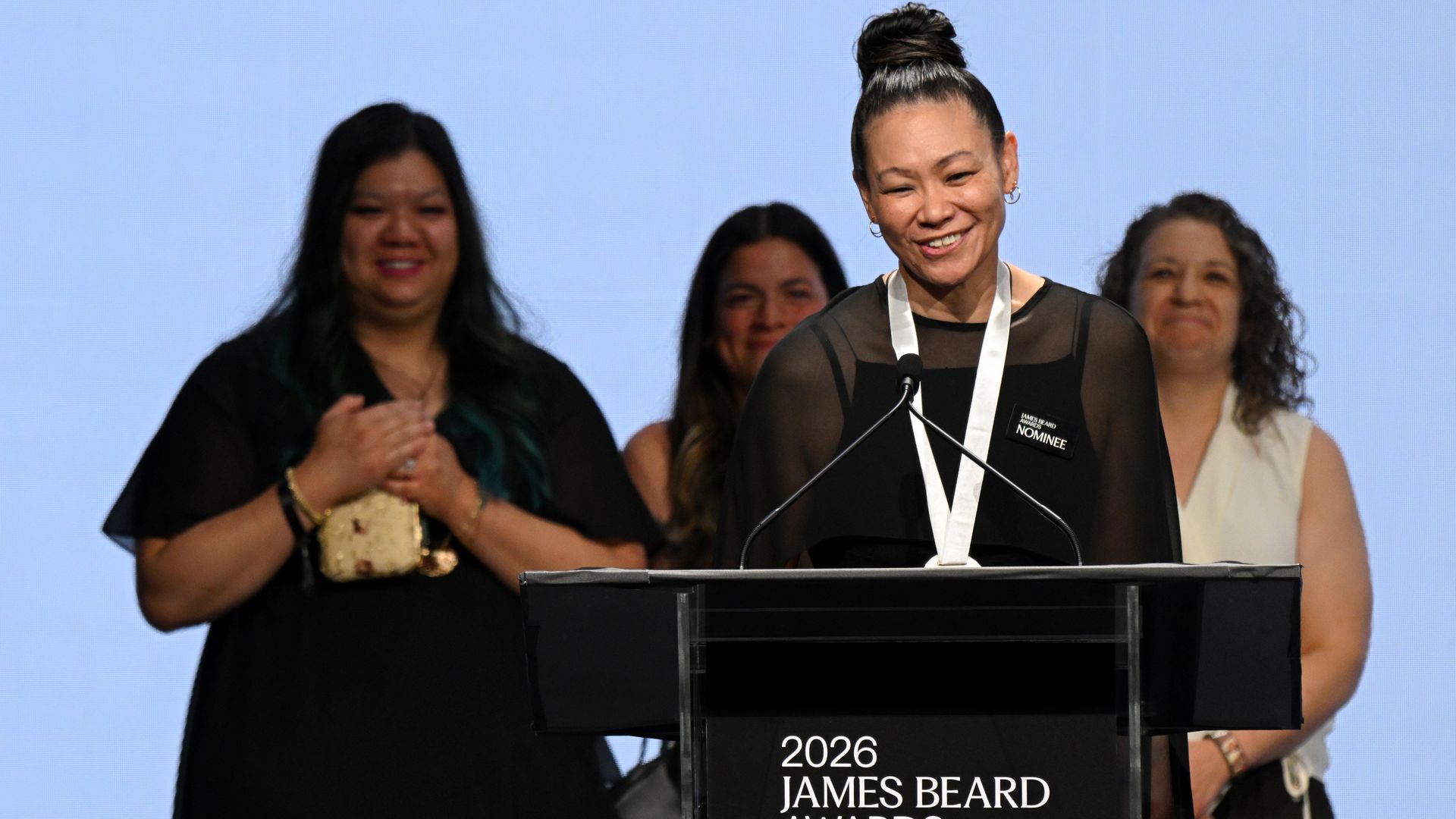 Smiling woman with a high bun standing at a black podium labeled "2026 JAMES BEARD AWARDS" speaks into a microphone, while people applaud behind her against a blue sky backdrop.