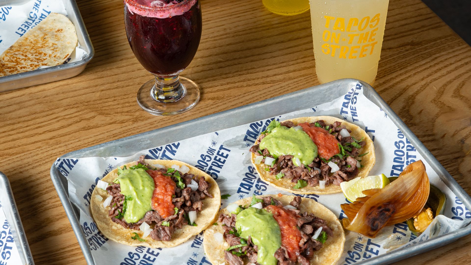 Three tacos with beef, green and red sauce on a tray with grilled onion and lime wedge on a wooden table, alongside three drinks including a pineapple Jarritos soda.
