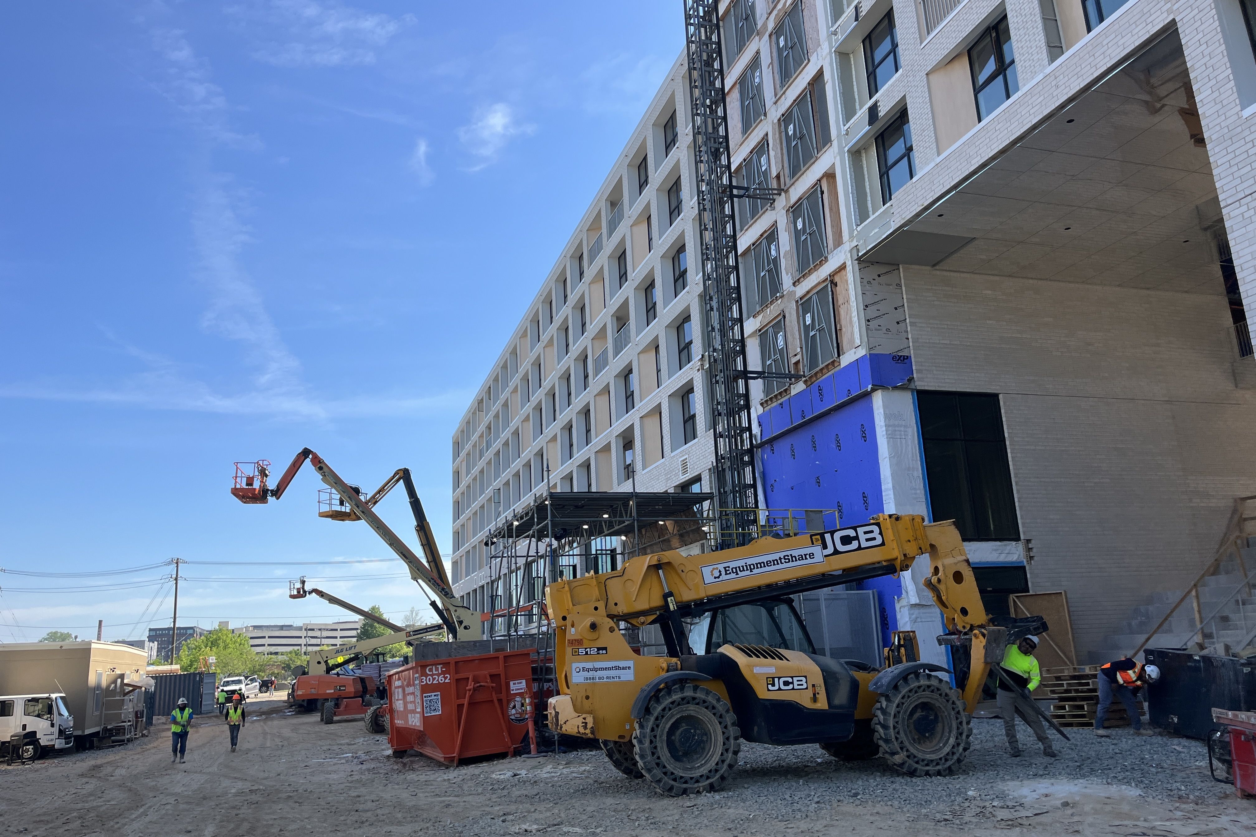 Construction site with a white brick building under development, metal scaffolding, and a yellow JCB crane. Workers in safety vests on a dirt road beneath a blue sky.