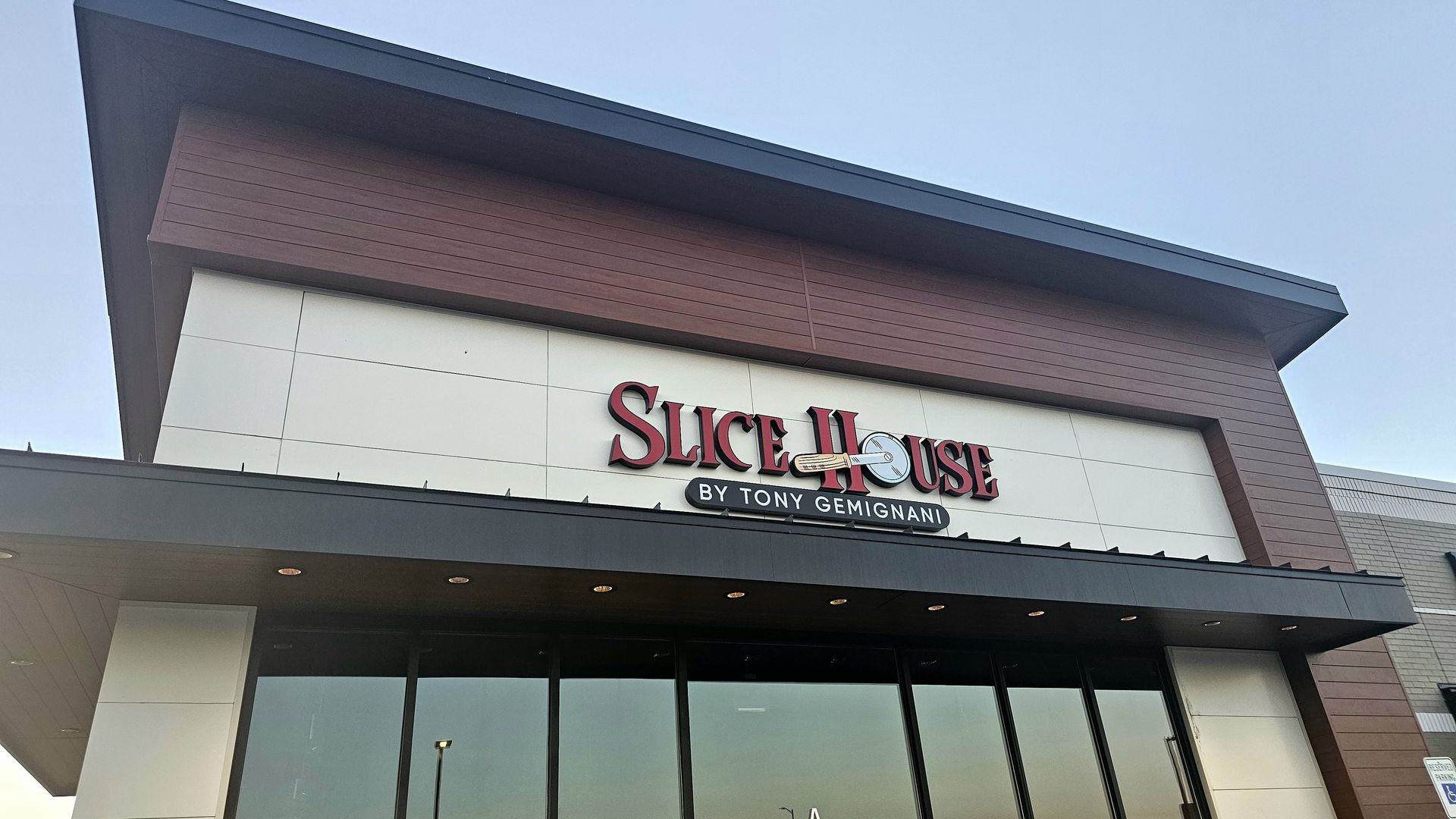 Exterior of Slice House by Tony Gemignani, a modern storefront with large glass windows, brown horizontal siding above, and a red-and-black sign featuring a pizza image.