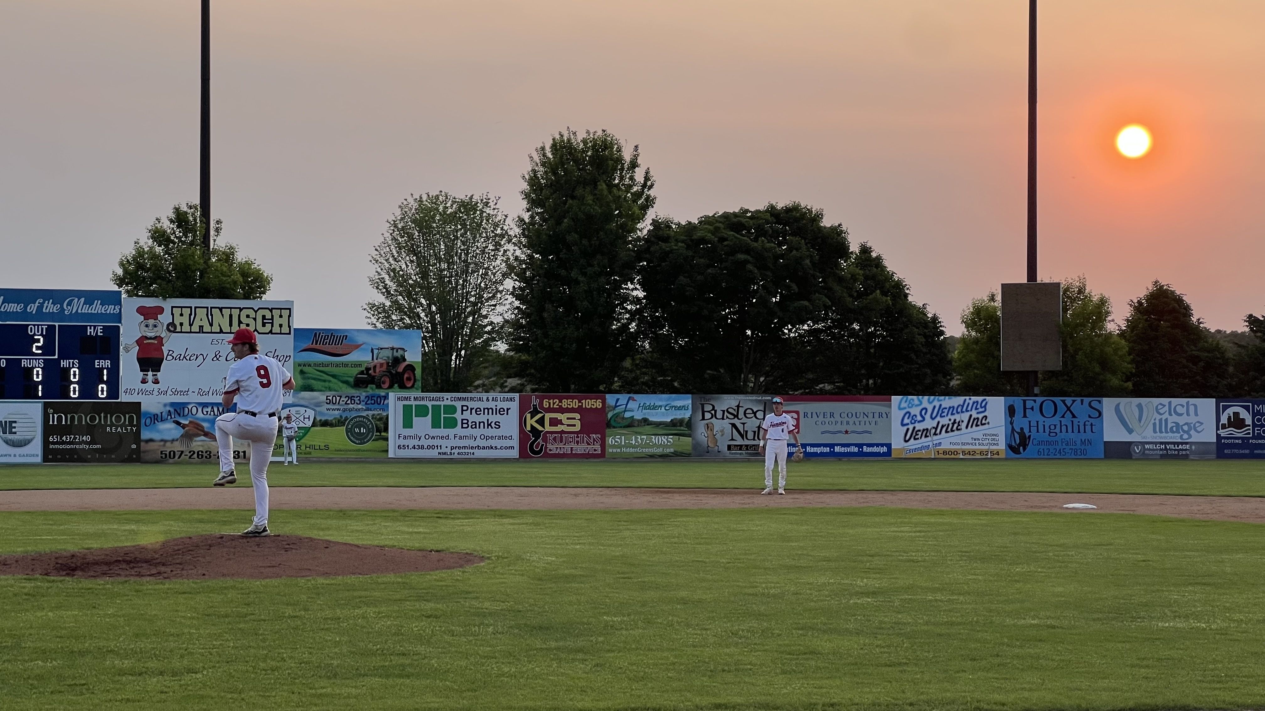 A baseball pitcher on the mound delivers a pitch as the sun sets behind him