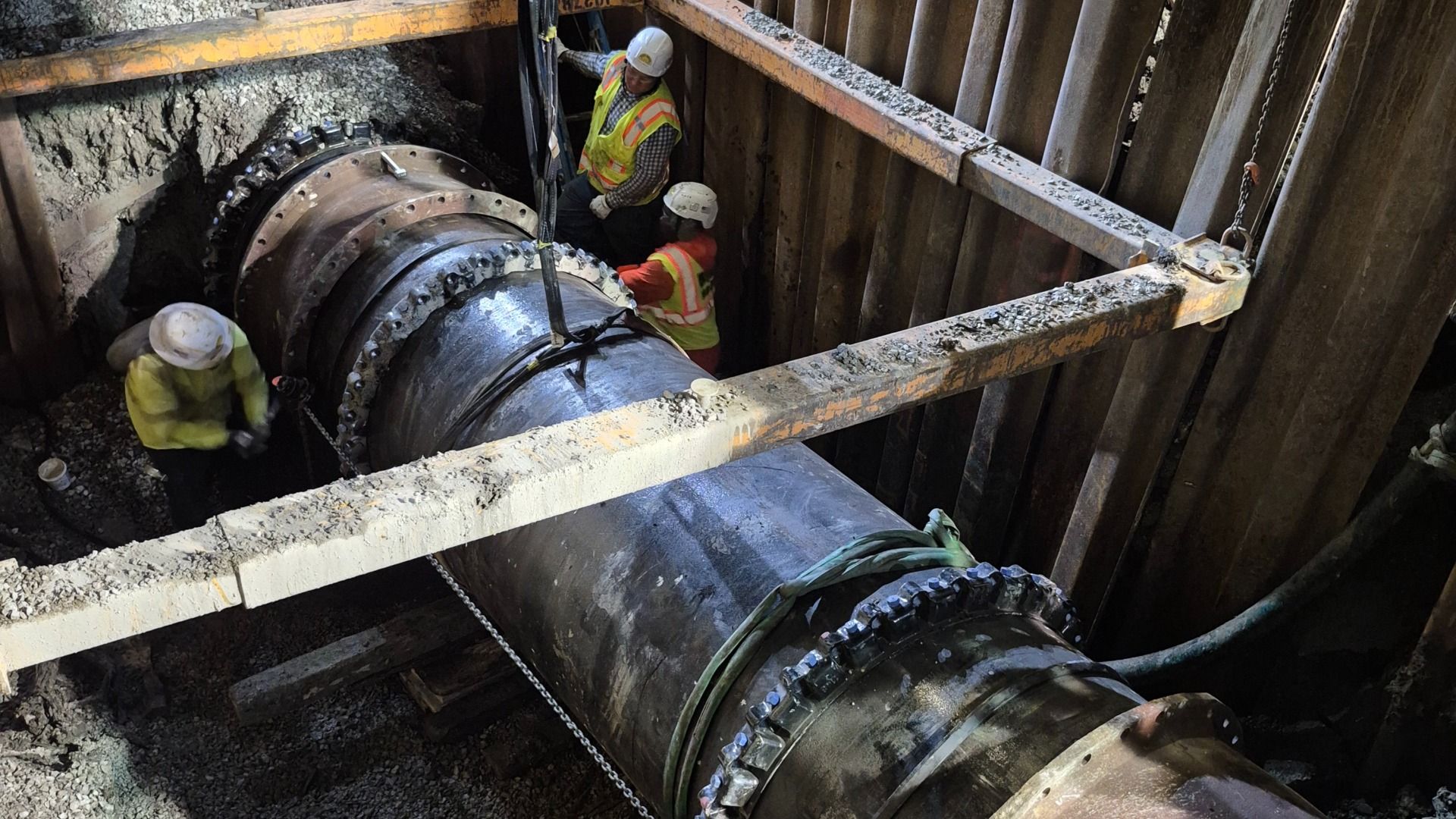 Workers in hard hats and hi-vis vests in a trench, guiding a large bolted steel pipe with flanges; muddy walls and steel shoring surround the excavation.