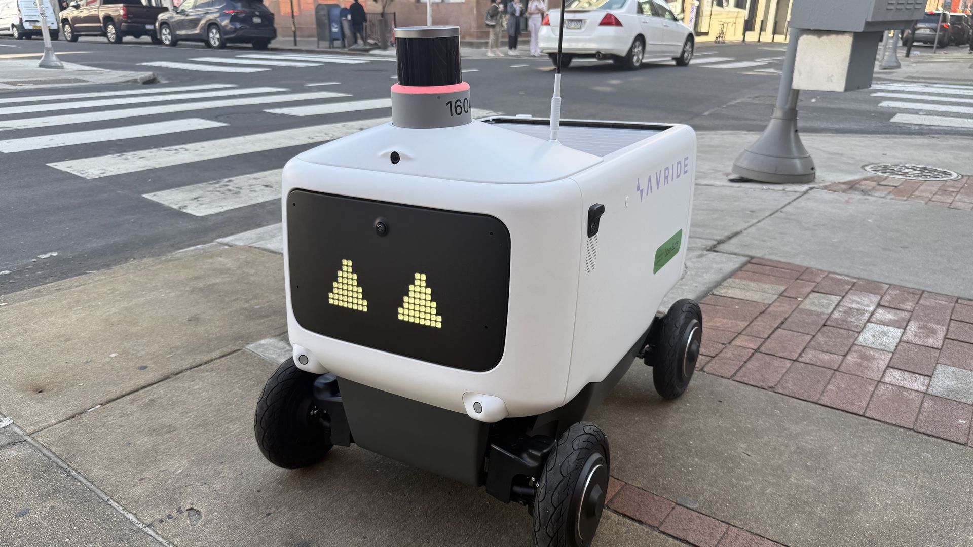 White autonomous delivery robot with a black front display showing two yellow pixel triangles, four wheels and a pink ring on top, on a city sidewalk near a crosswalk and storefronts.
