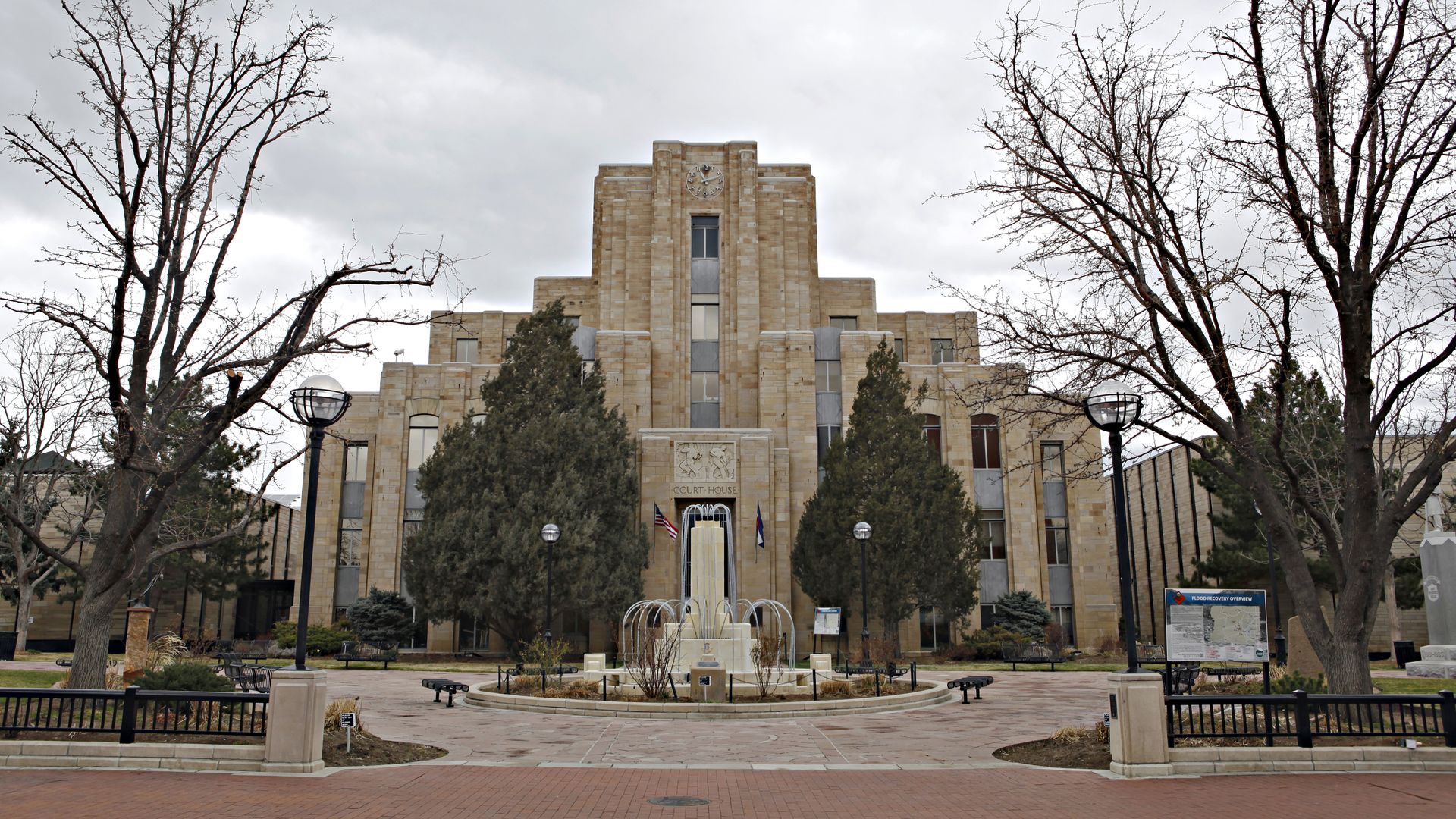 Art Deco courthouse building with a clock on top, surrounded by bare trees, evergreen shrubs, and a central fountain in front under a cloudy sky.