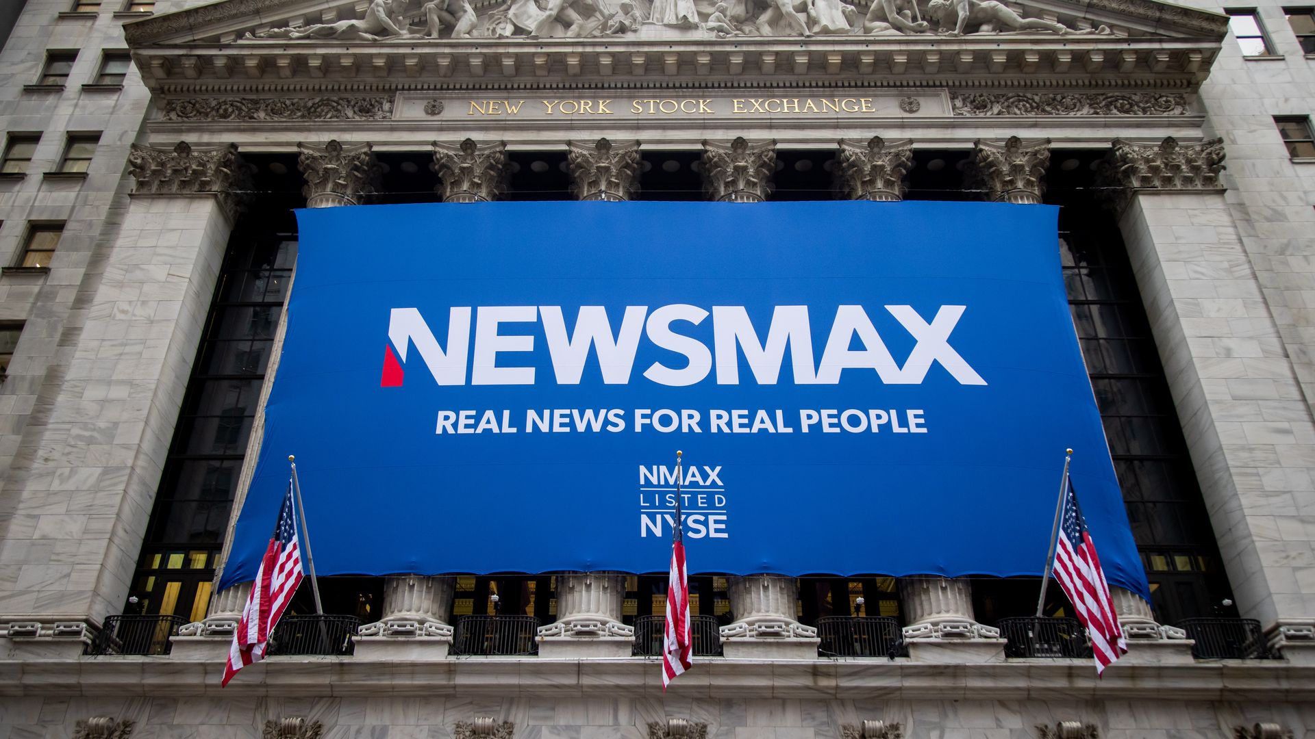 Blue NEWSMAX banner with slogan "REAL NEWS FOR REAL PEOPLE" hanging on New York Stock Exchange facade, which features classical columns and American flags.