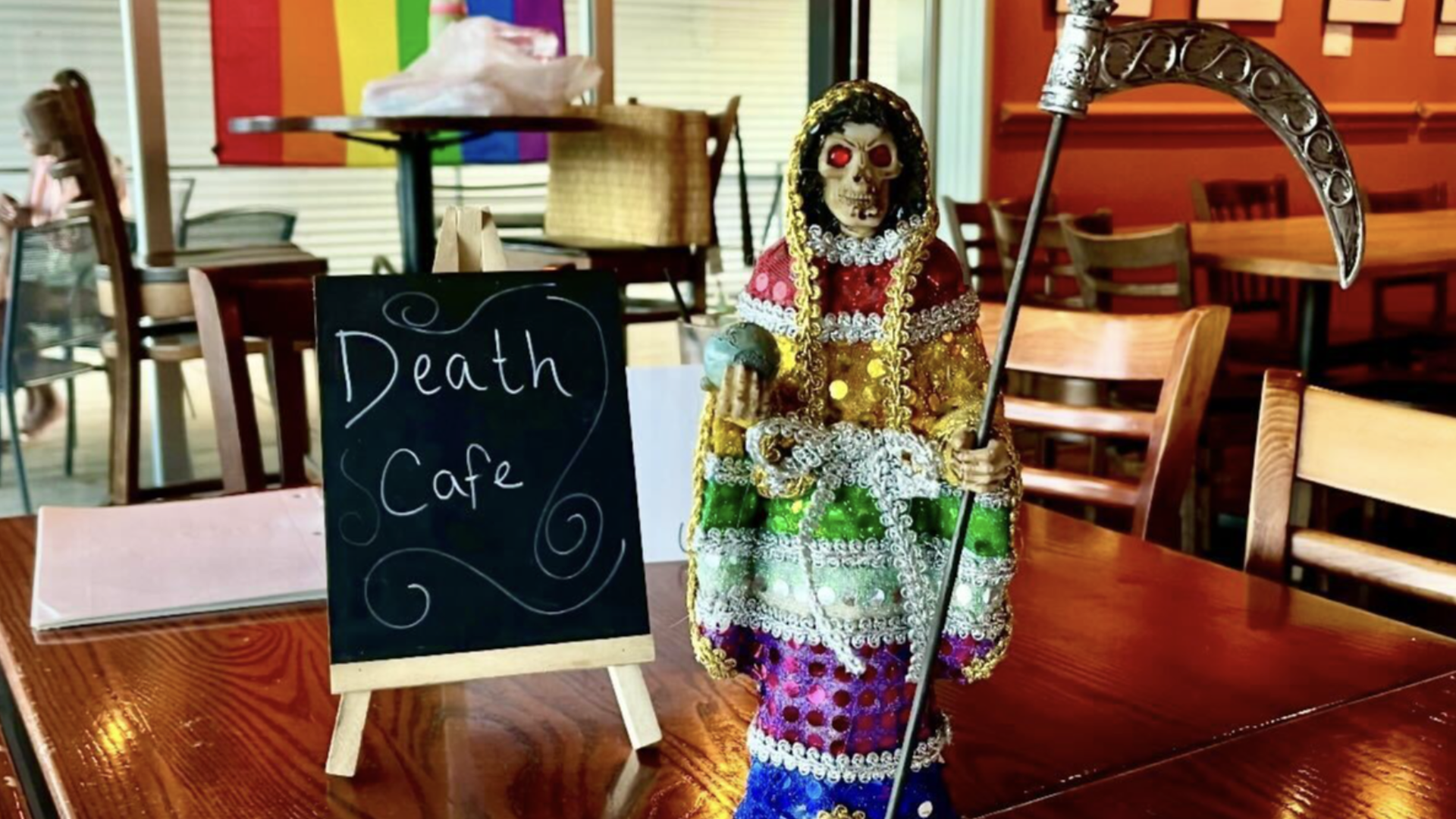 A colorful skeletal figurine holding a scythe and globe, dressed in multicolored sequins, stands next to a small blackboard sign that reads "Death Cafe" on a wooden table in a cafe.