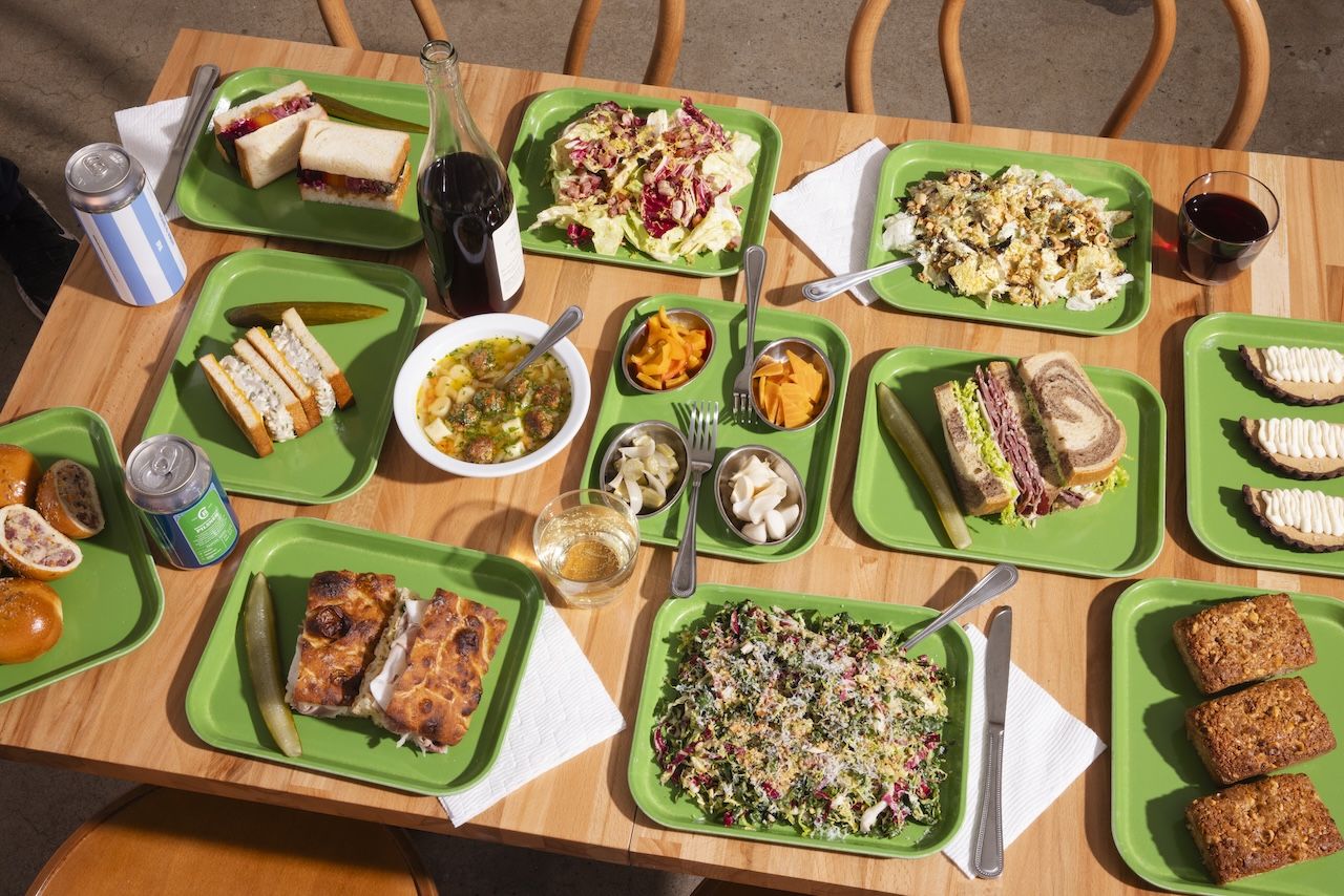 Wooden table with green trays holding various foods: sandwiches, salads, soup, pickles, small cups of pickled vegetables, dessert bars, and drinks in cans, bottles, and a glass.