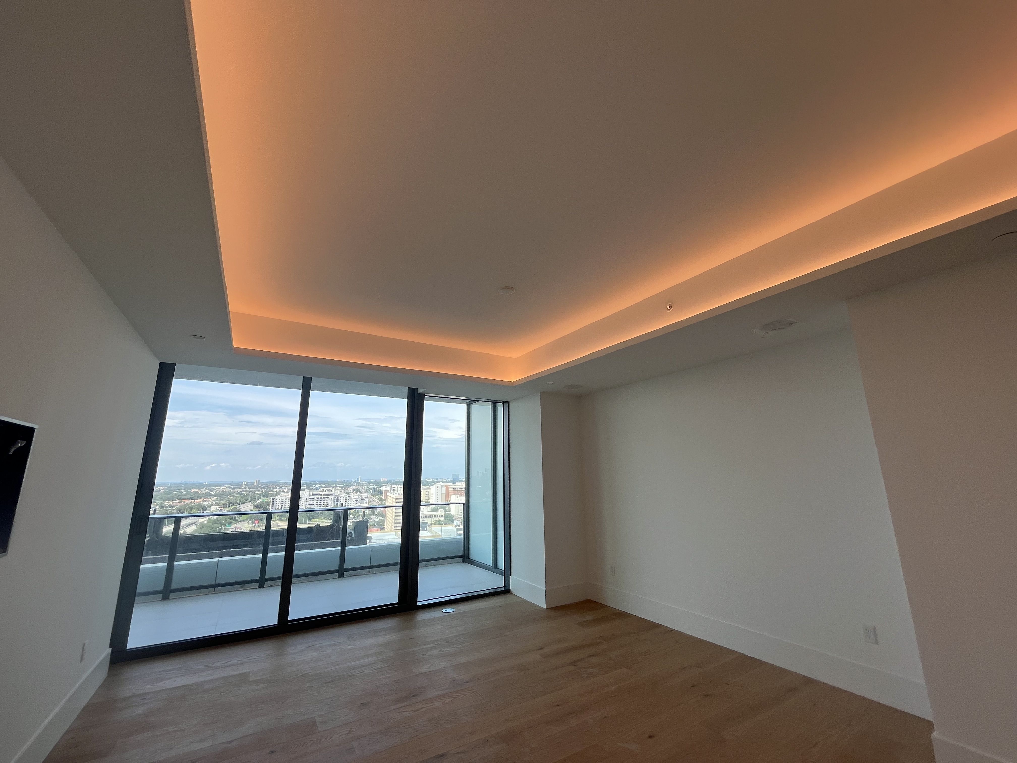 Modern living area with a recessed ceiling lit by warm orange LED lights. Floor-to-ceiling windows open to a balcony overlooking a cityscape; light wood floors and white walls.