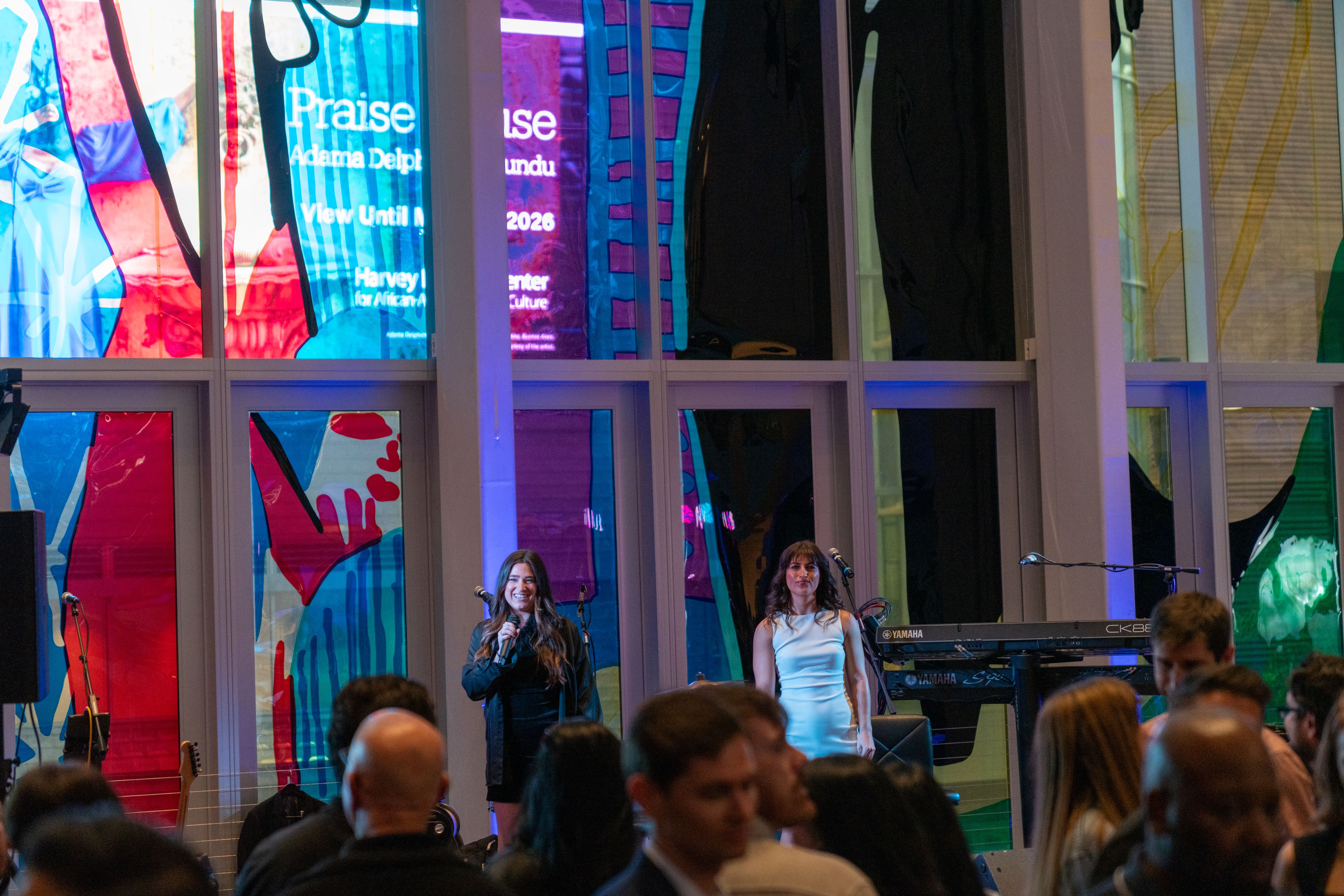 Two women on stage in front of a crowd, one speaking into a microphone, with colorful abstract artwork on the windows behind them and Yamaha keyboard on the right side.