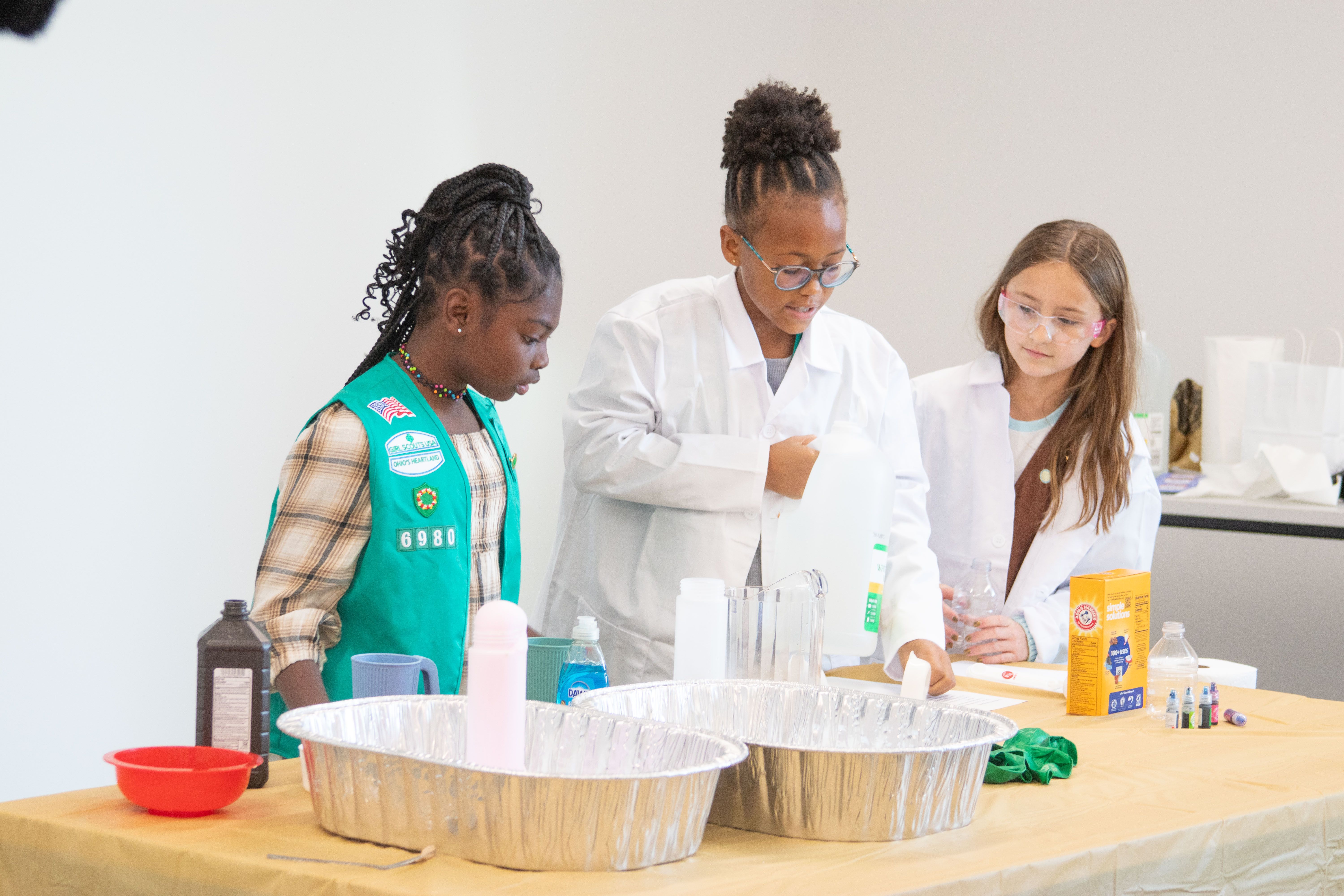 Three Girl Scouts, two in white robes and one in a vest, experiment with liquids and baking soda in a lab