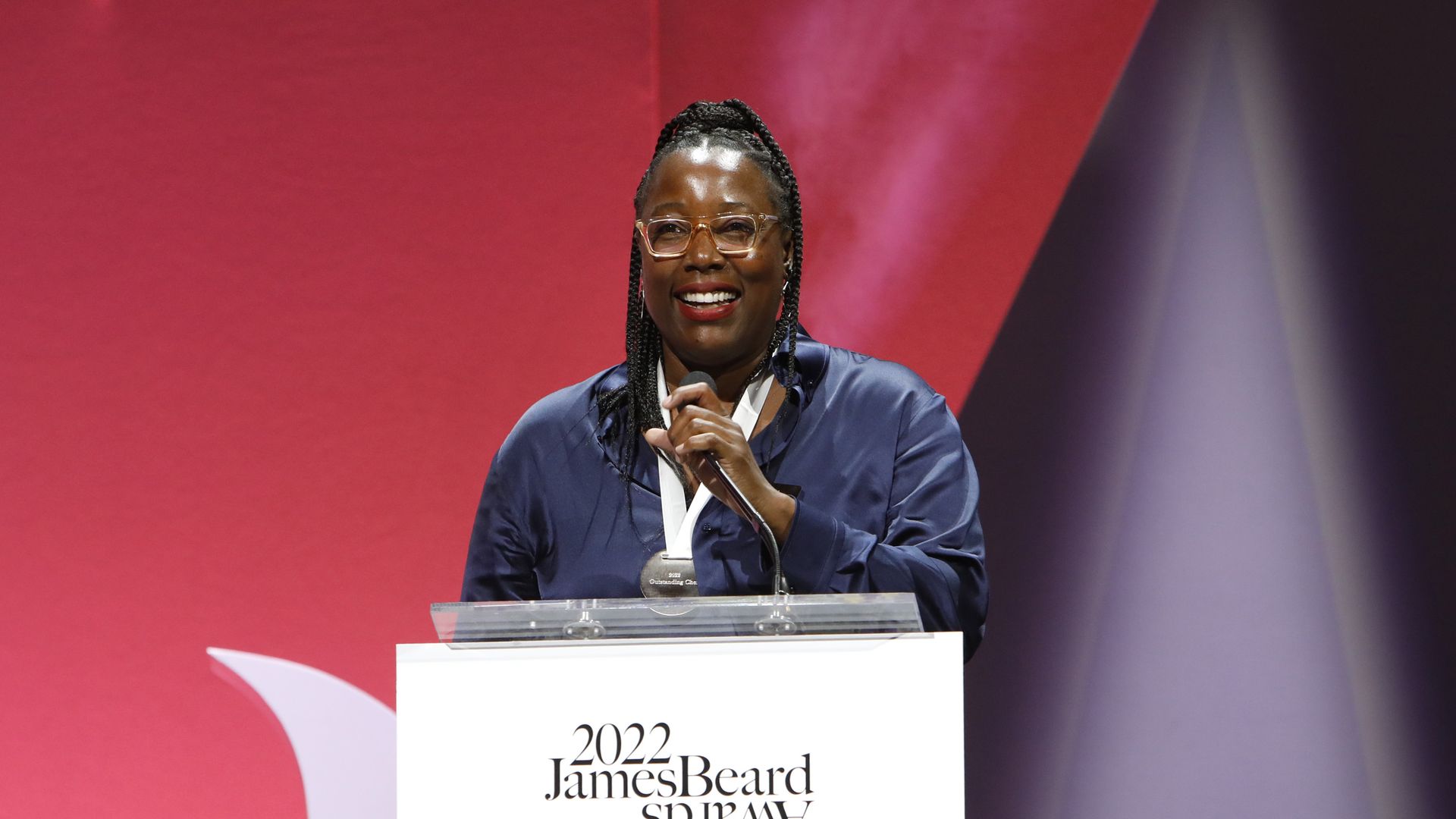 A Black woman with glasses and red lipstick smiles as she stands behind a podium speaking 