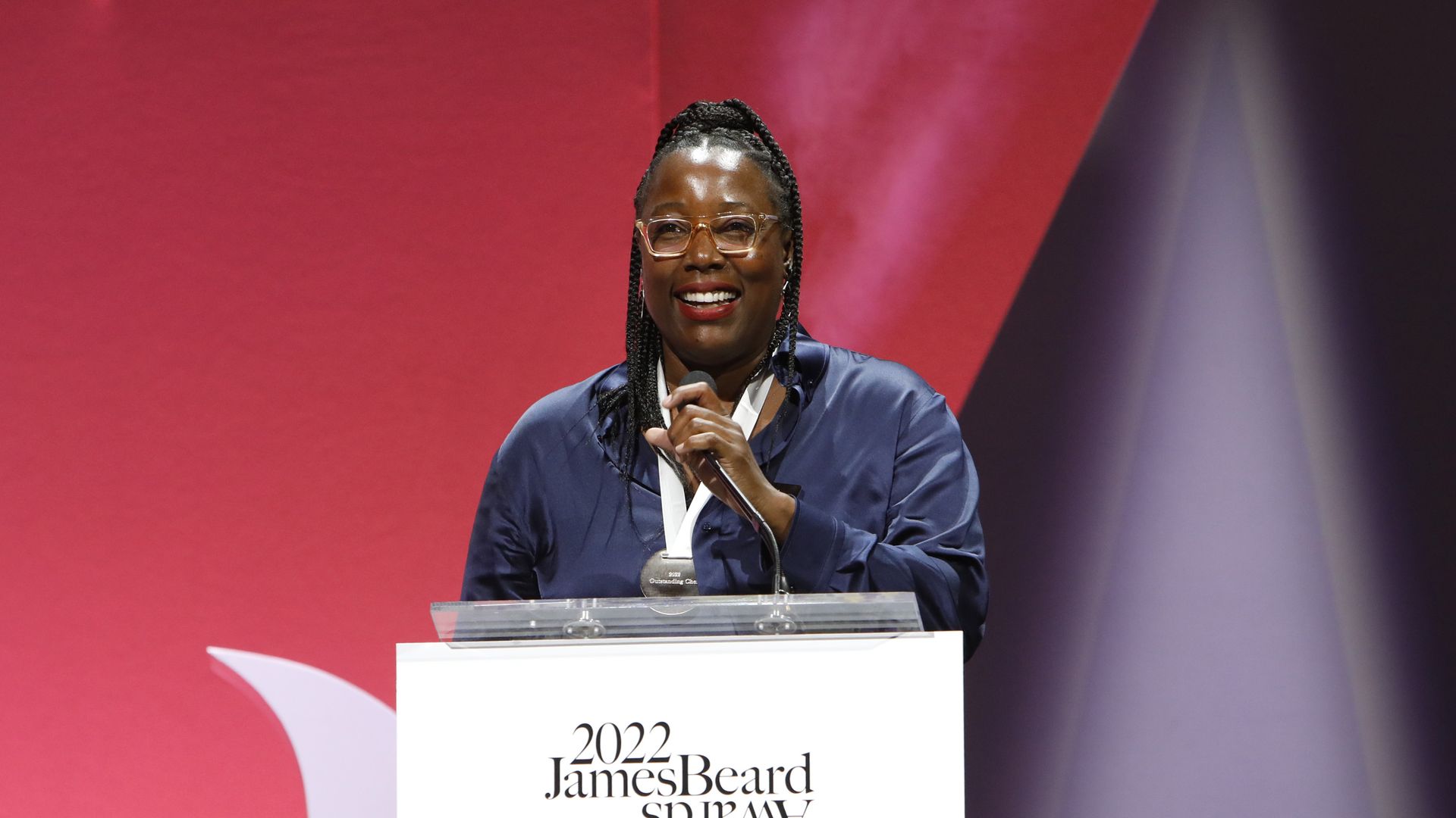 A Black woman with glasses and red lipstick smiles as she stands behind a podium speaking