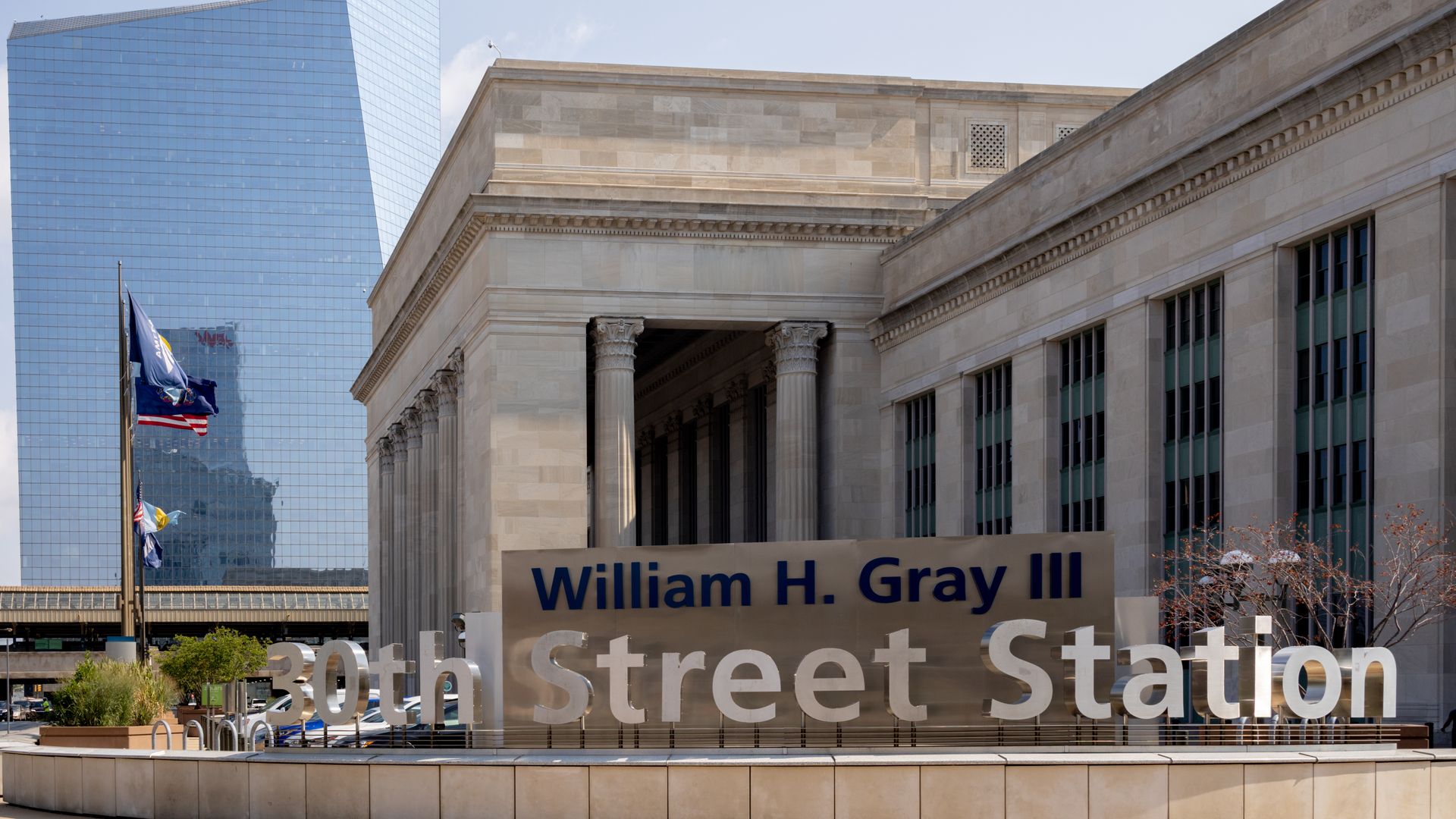 City scene with a tall blue glass skyscraper on the left and a beige neoclassical building with columns on the right. Flags fly; a large banner reads "William H. Gray III" and "30th Street Station."