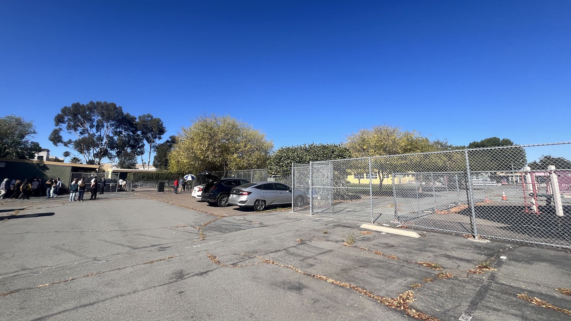 Outdoor parking lot with three cars along a metal fence enclosing a playground with pink structures and orange cones, people line up near a building under clear blue sky.