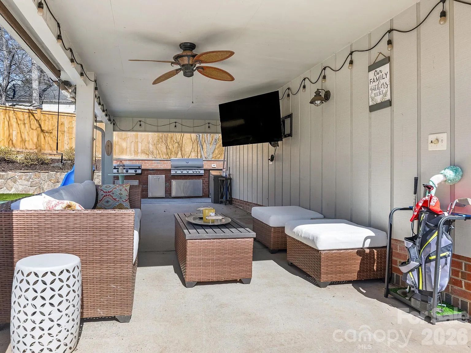 Covered patio with brown wicker furniture including sofa and stools with white cushions, ceiling fan, wall-mounted TV, string lights, outdoor grill, and golf bag with clubs.