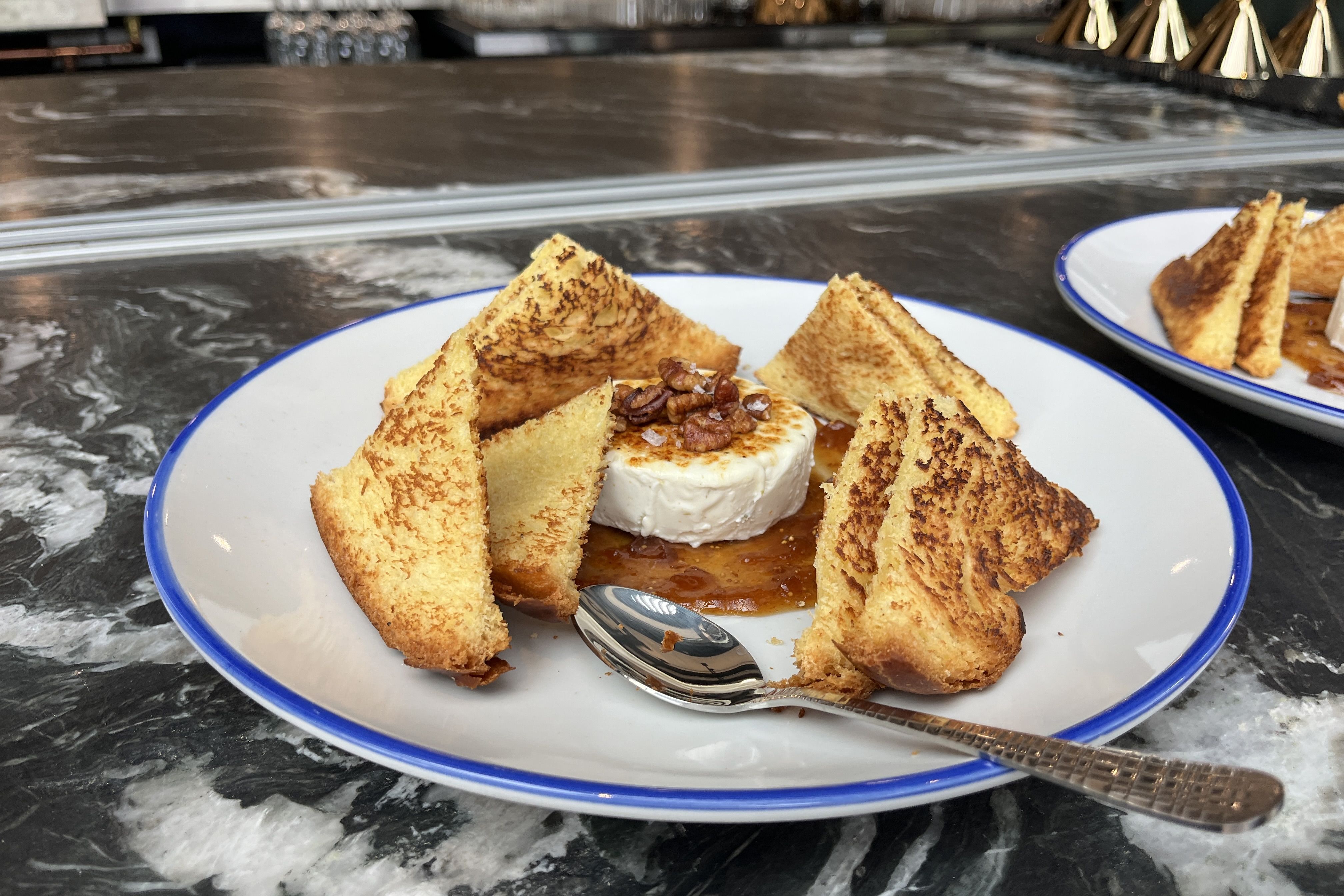Plate with a round brie in caramel sauce, surrounded by golden-brown triangular toasts; white plate with a blue rim on a dark marble counter, with another plate visible in the background.