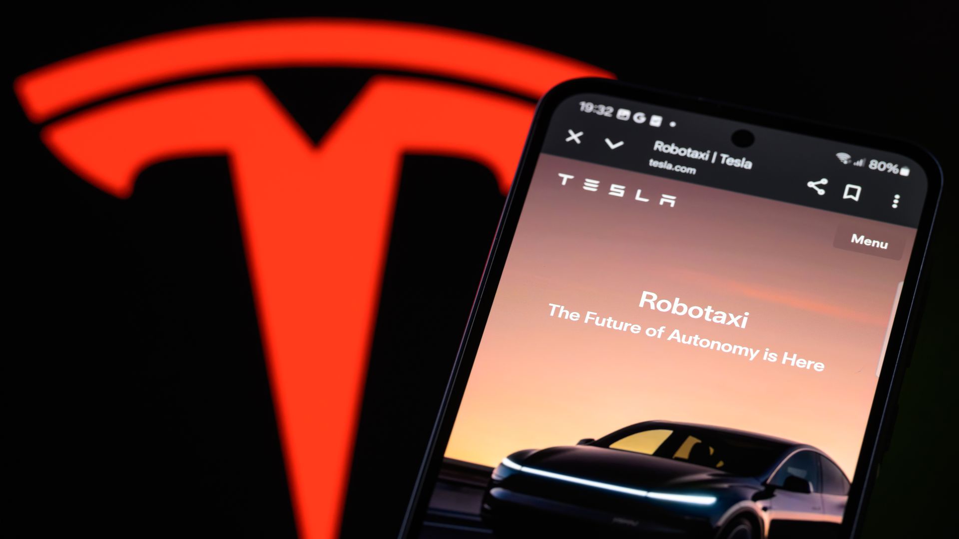 Close up of a phone screen showing Tesla's Robotaxi app, including a photo of a dark car, with thin light bar on across the front, and orange sunset. Also shown a red Tesla T-shaped logo. 
