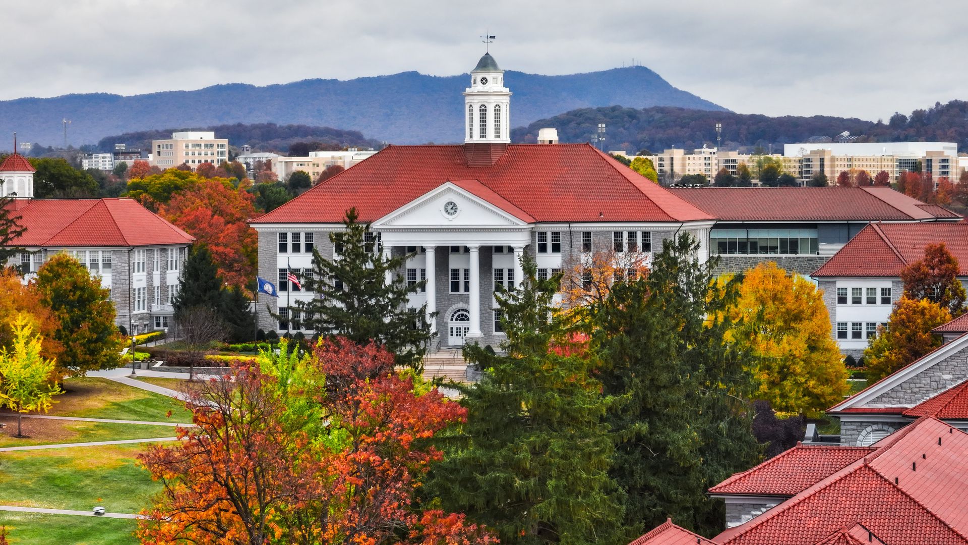 Campus building with a white clock tower and red-tiled roof, surrounded by autumn trees in orange, yellow, and green; mountains and cloudy sky in the distance.