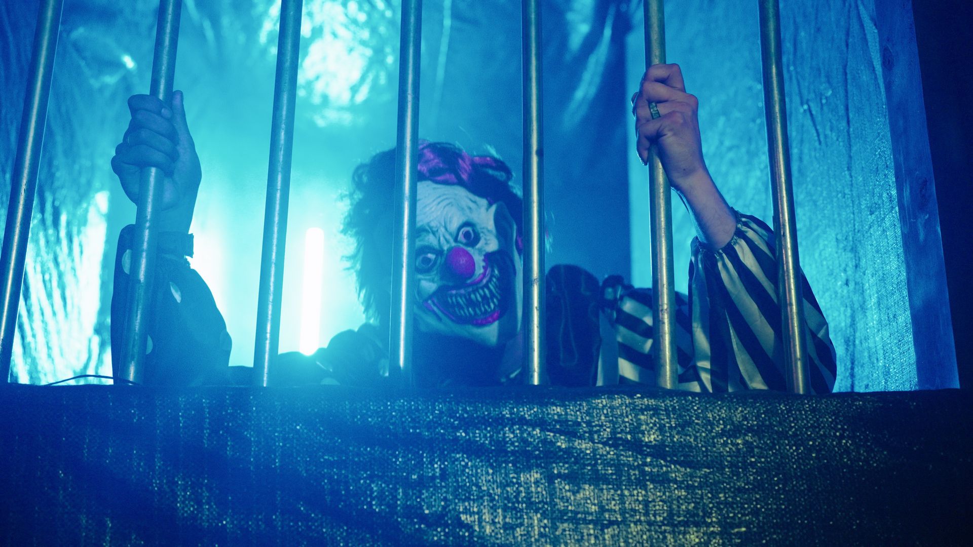 Clown with white makeup and a red nose grips vertical bars in a blue-lit, jail-like scene; striped sleeves and a menacing expression show through the bars.