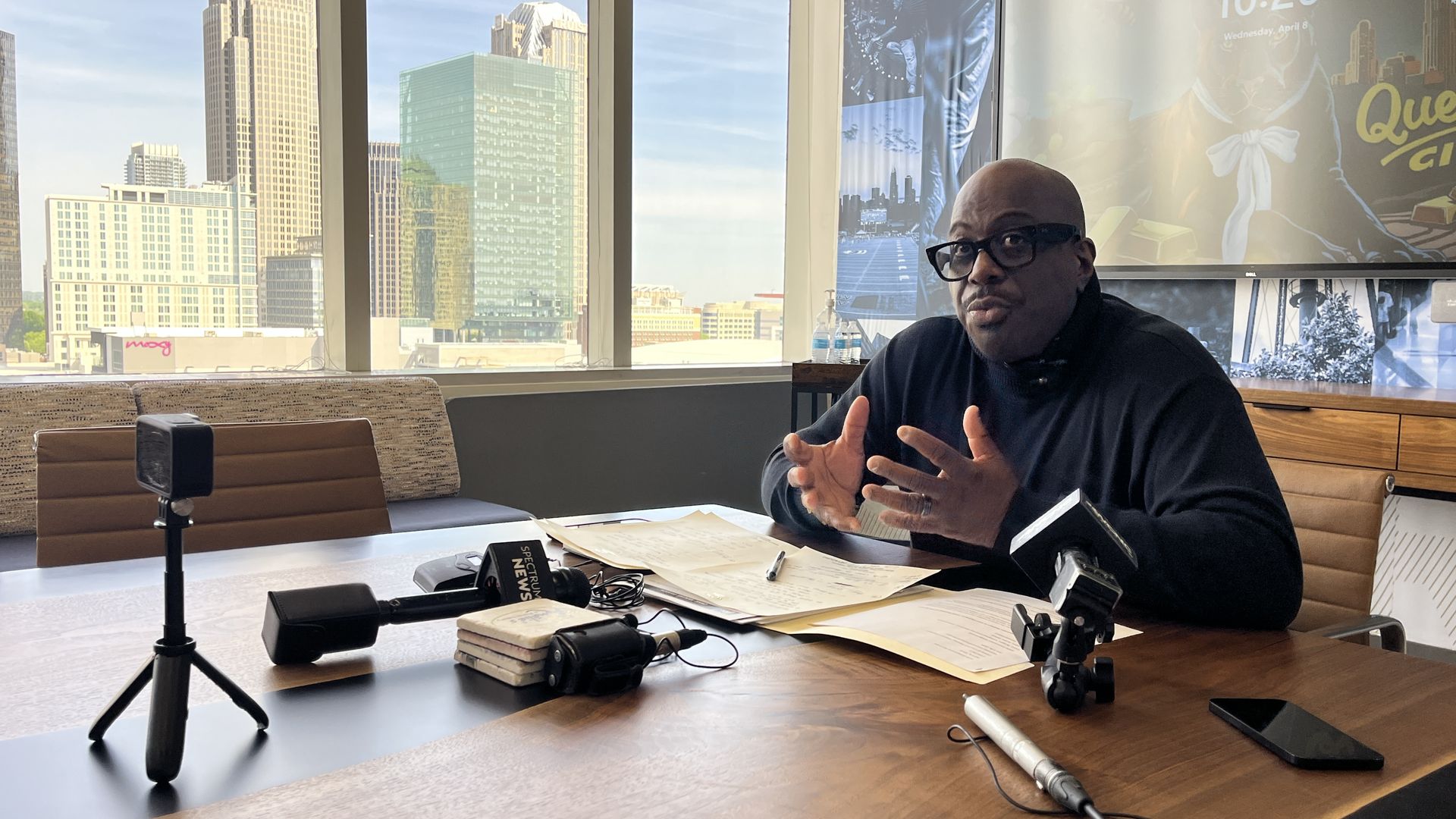 A man in a black sweater and glasses sits at a wooden conference table with papers and microphones; city skyline outside large windows and a screen showing 10:20 behind him.