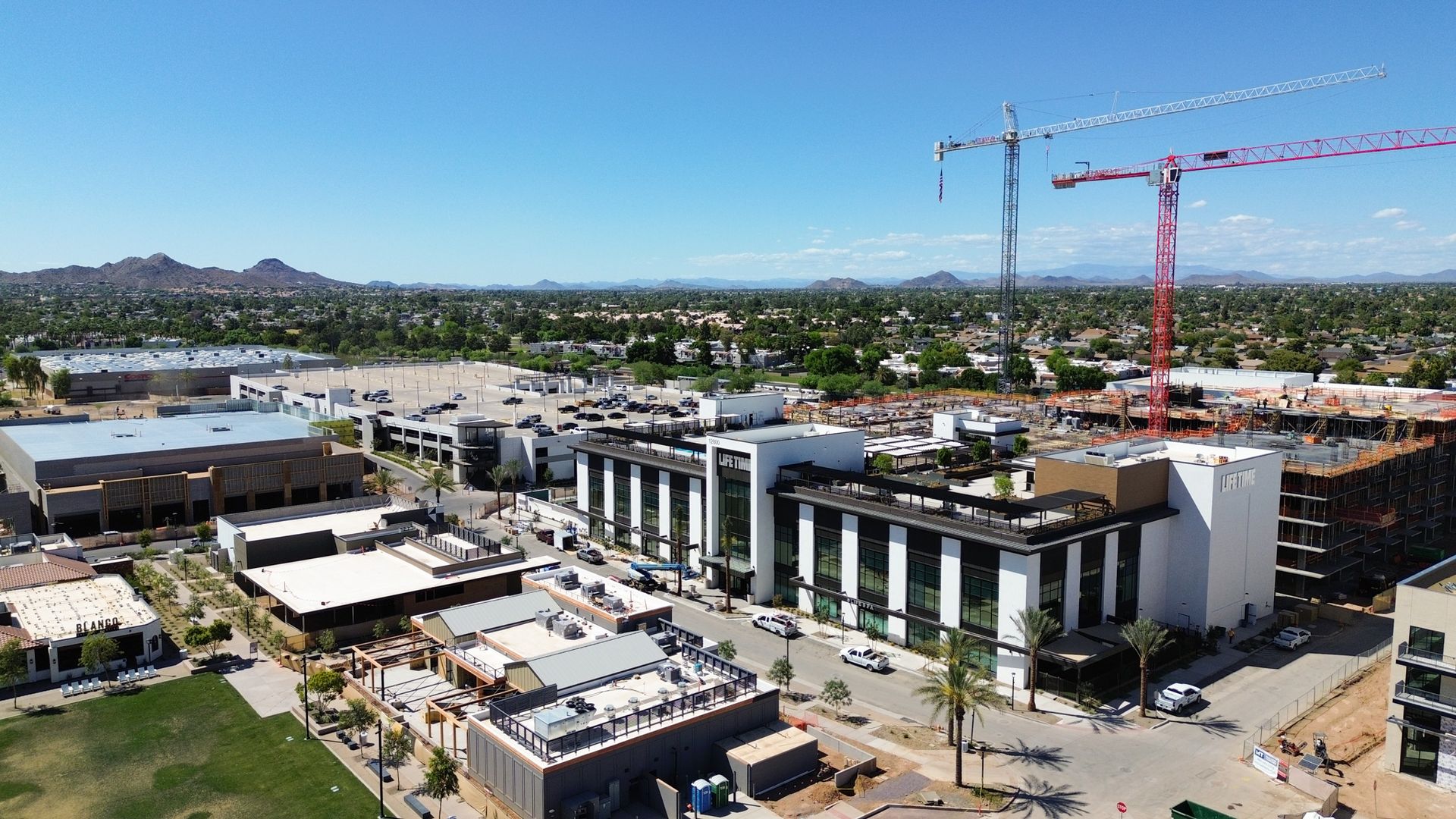 Clear blue sky over a developing commercial complex: two tall construction cranes, one red and one gray, rise above modern white buildings; a parking lot and distant desert mountains complete the view.