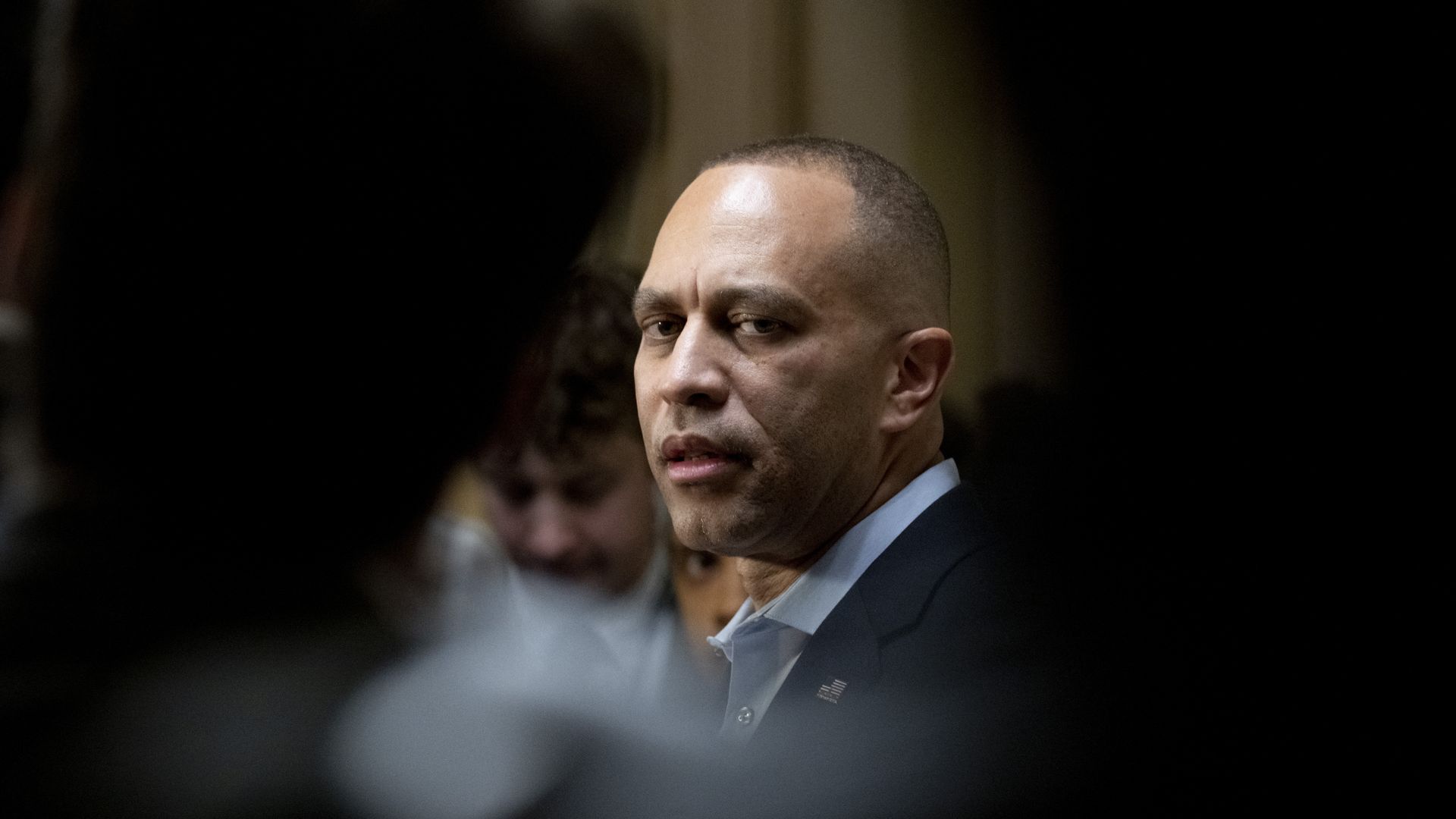 House Minority Leader Hakeem Jeffries, wearing a tieless blue suit and seen through the silhouettes of two reporters in a yellowish-beige hallway.